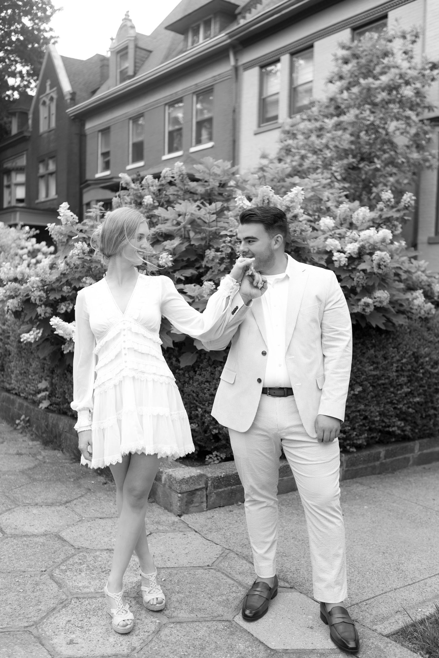 Black and white photo of a couple in white outfits posing in front of flowering bushes and brick buildings.