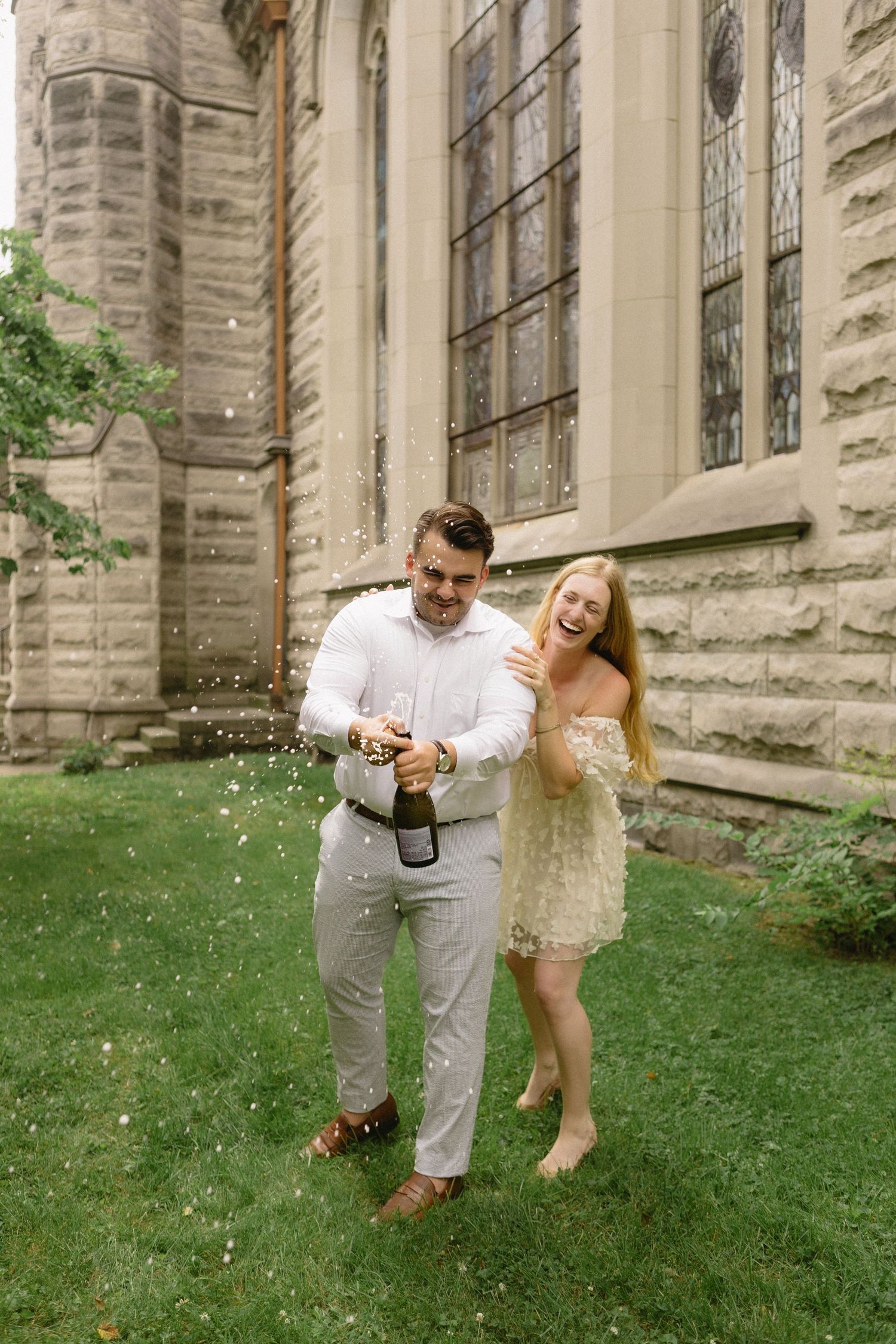 Couple popping champagne bottle outside historic stone church, woman in lace dress laughing joyfully during celebration.