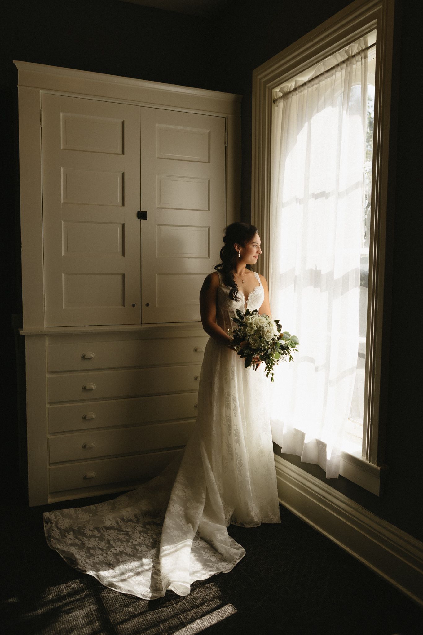 A bride in a flowing white dress holds a bouquet while standing in dramatic window light next to a built-in wardrobe.