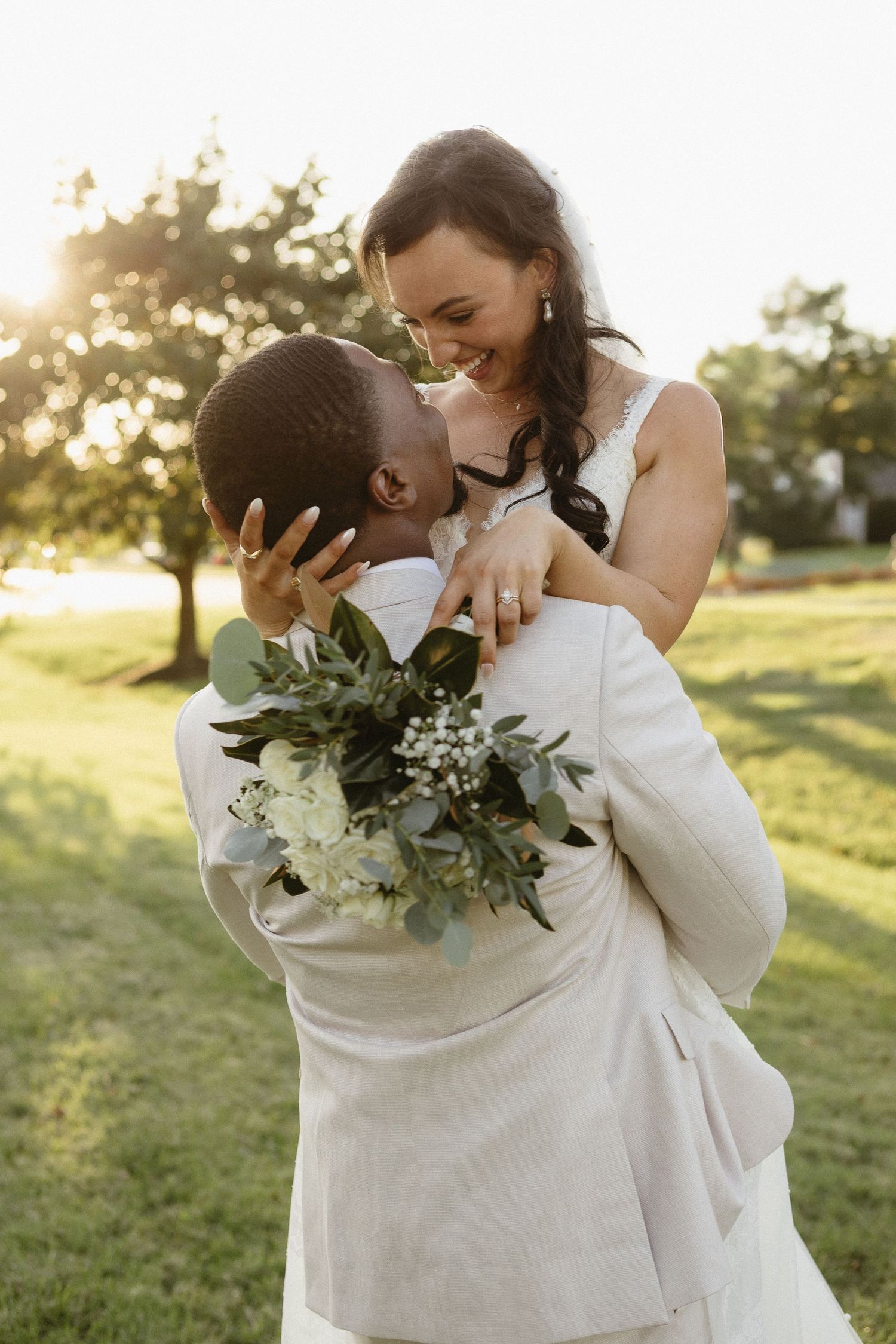 A joyful wedding couple embraces outdoors at sunset with a beautiful floral bouquet.
