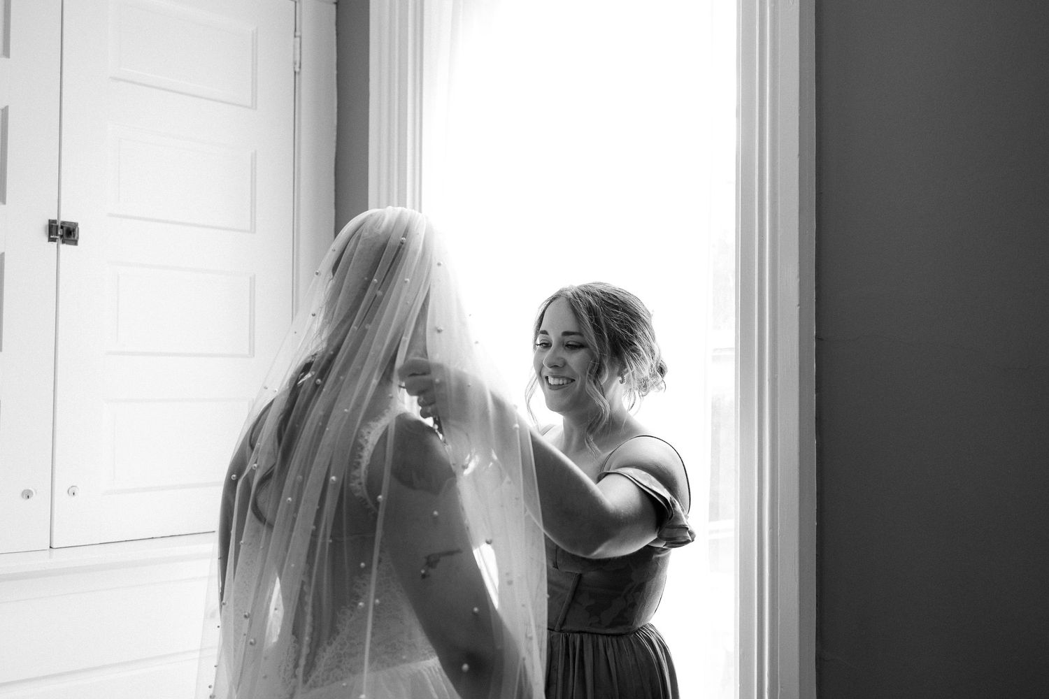 Black and white photo of a tender moment during wedding preparations near a window.