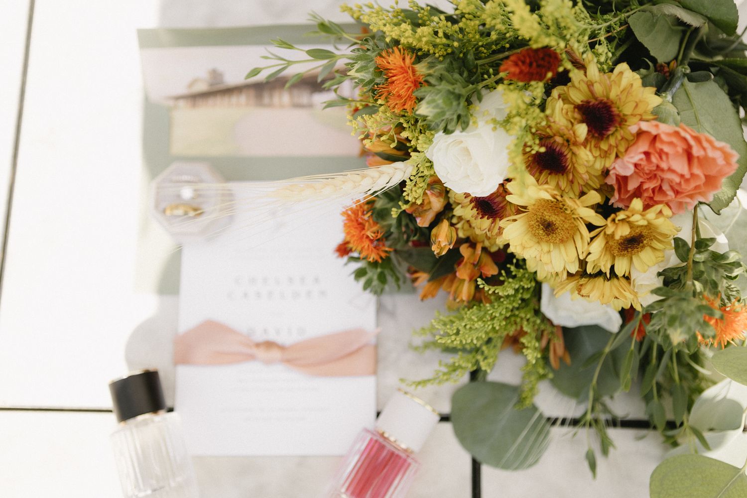 Vibrant floral arrangement with sunflowers, peach roses and greenery next to wedding stationery on white surface.