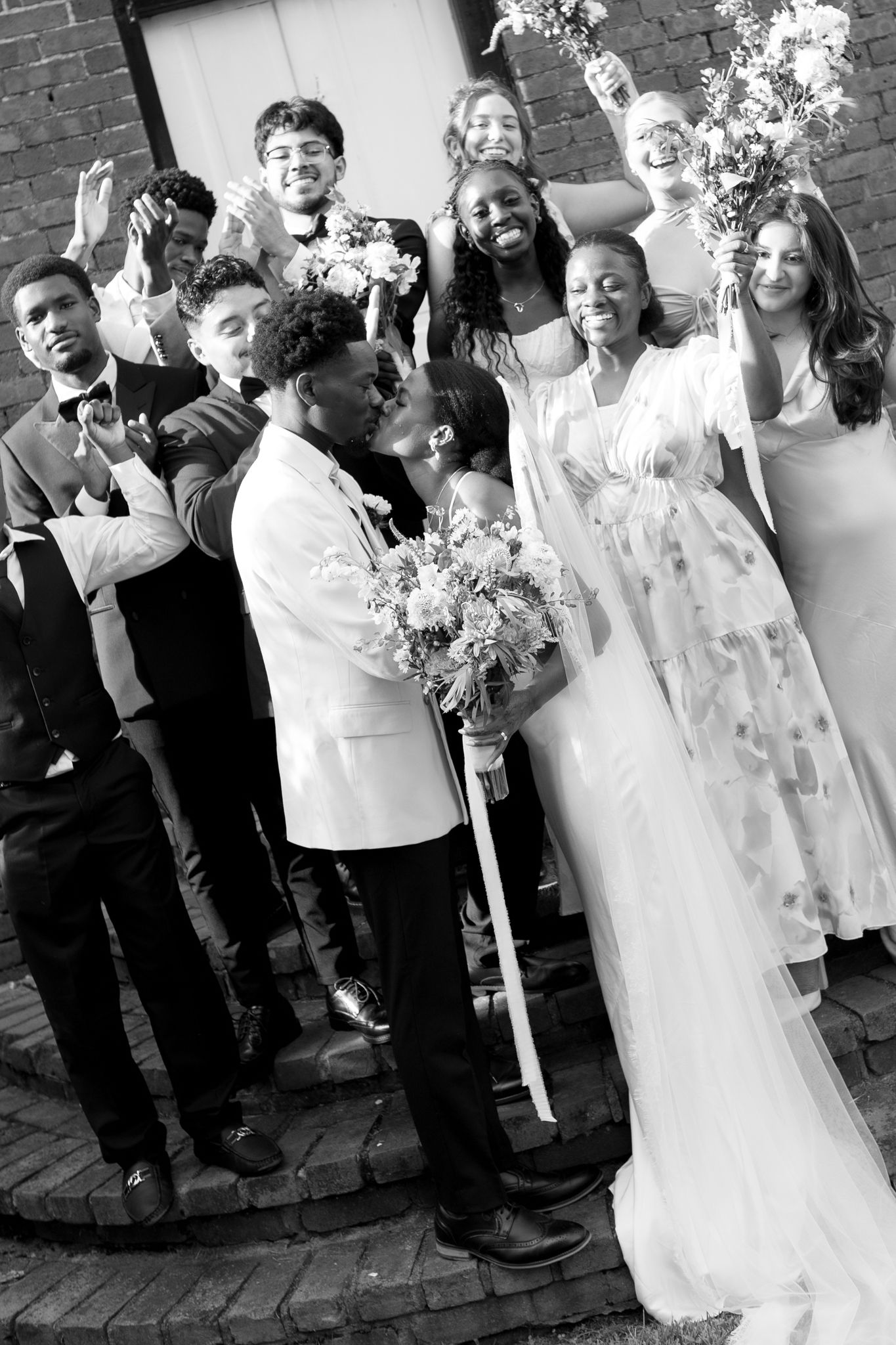 Wedding party celebrates joyfully against brick wall backdrop in black and white photography.