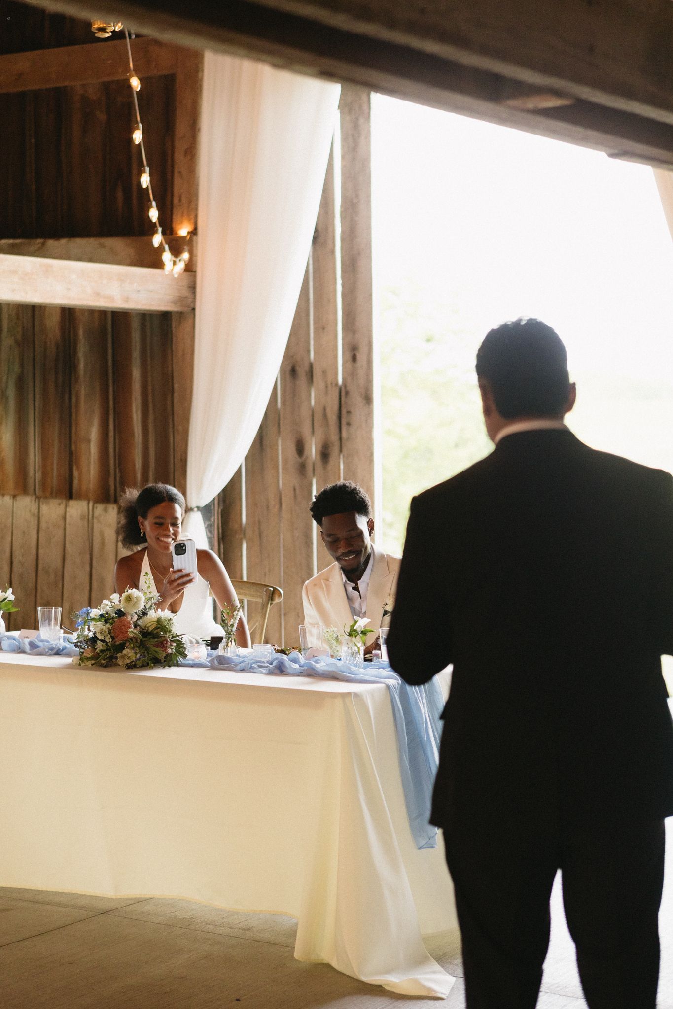 A reception scene in a rustic barn venue with string lights and white table linens.