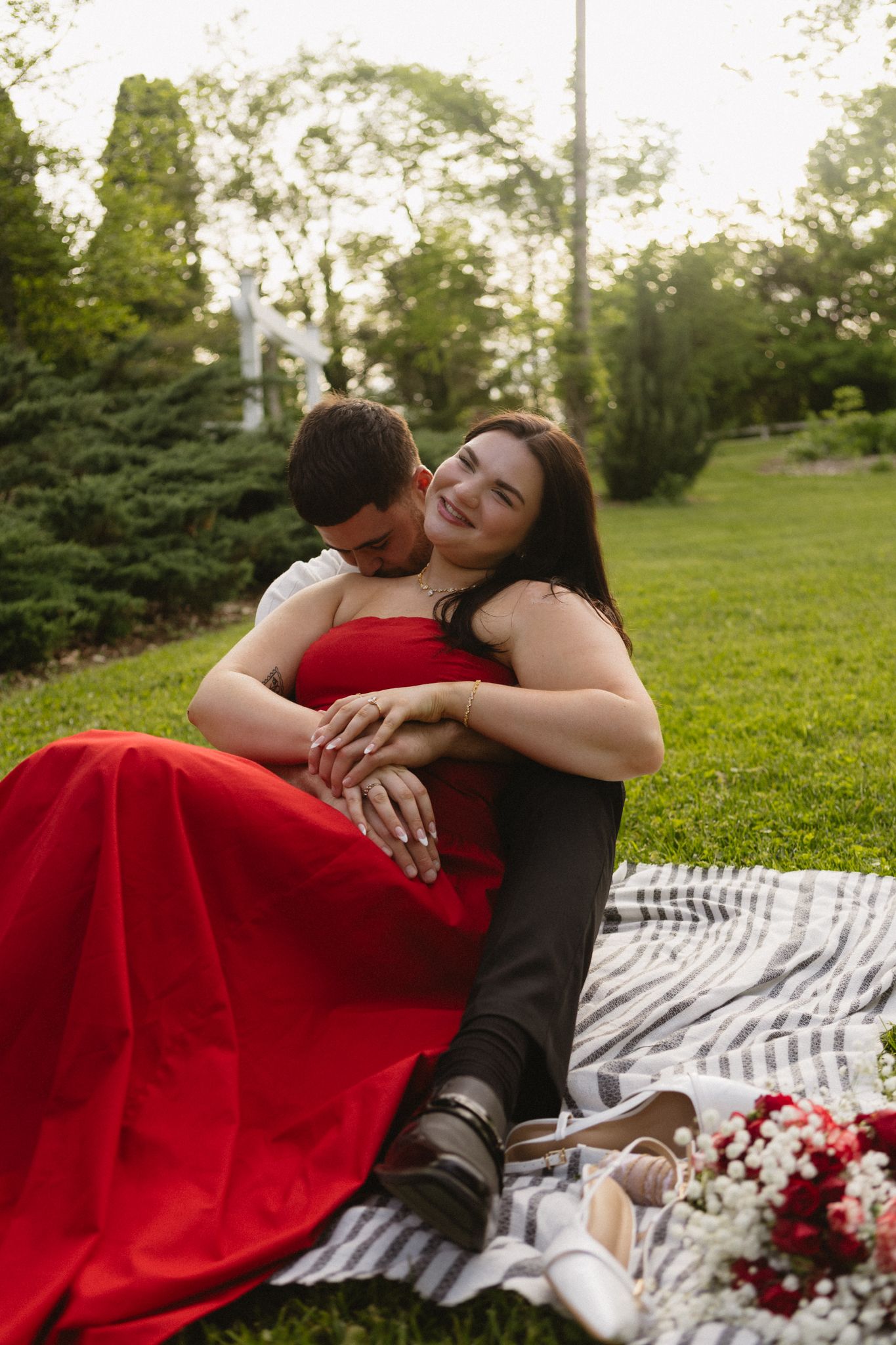 A couple shares a tender moment on a striped blanket surrounded by scattered red rose petals in a sunlit park.