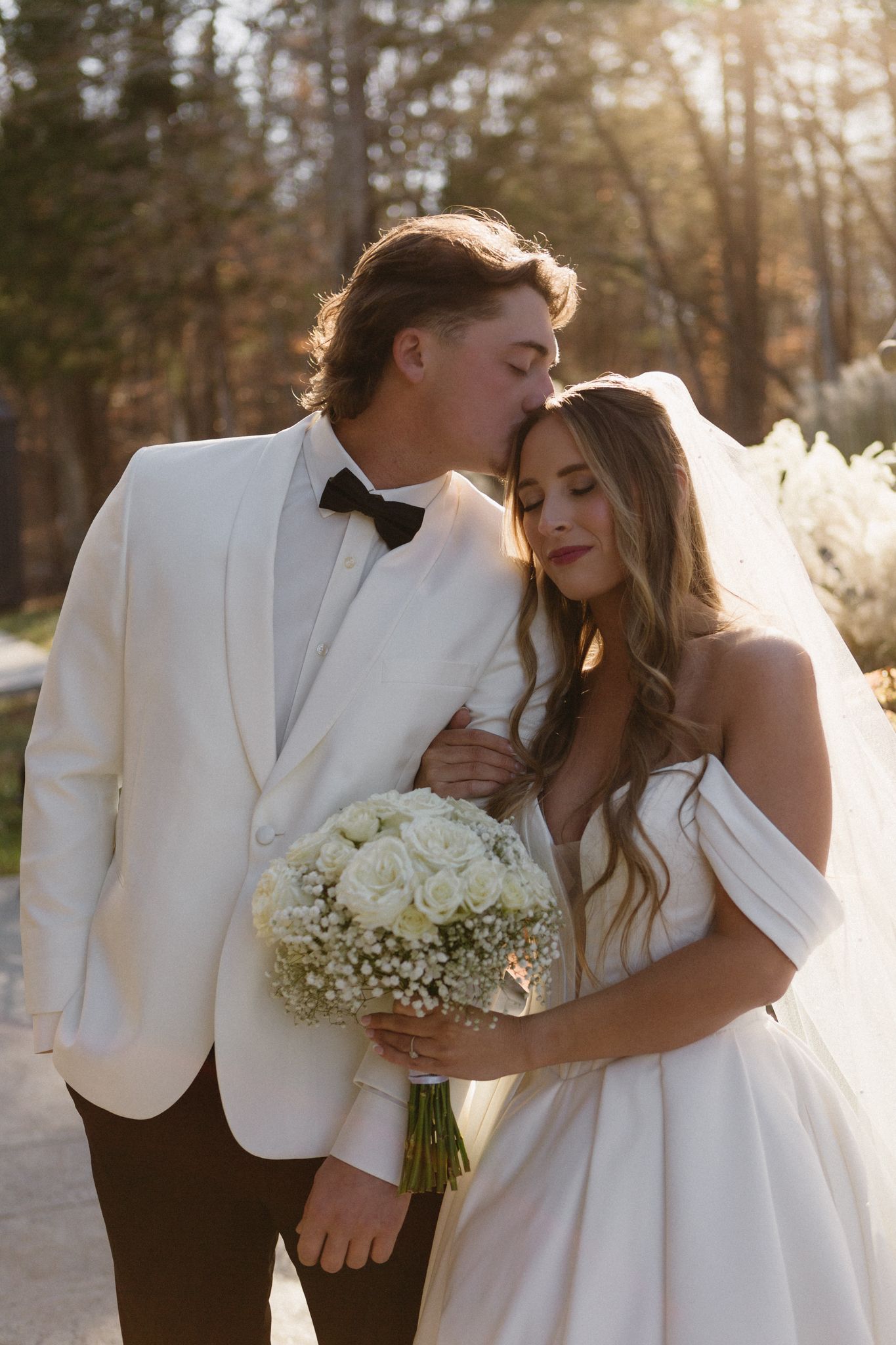 A wedding couple embraces in romantic sunset lighting while holding a white floral bouquet.