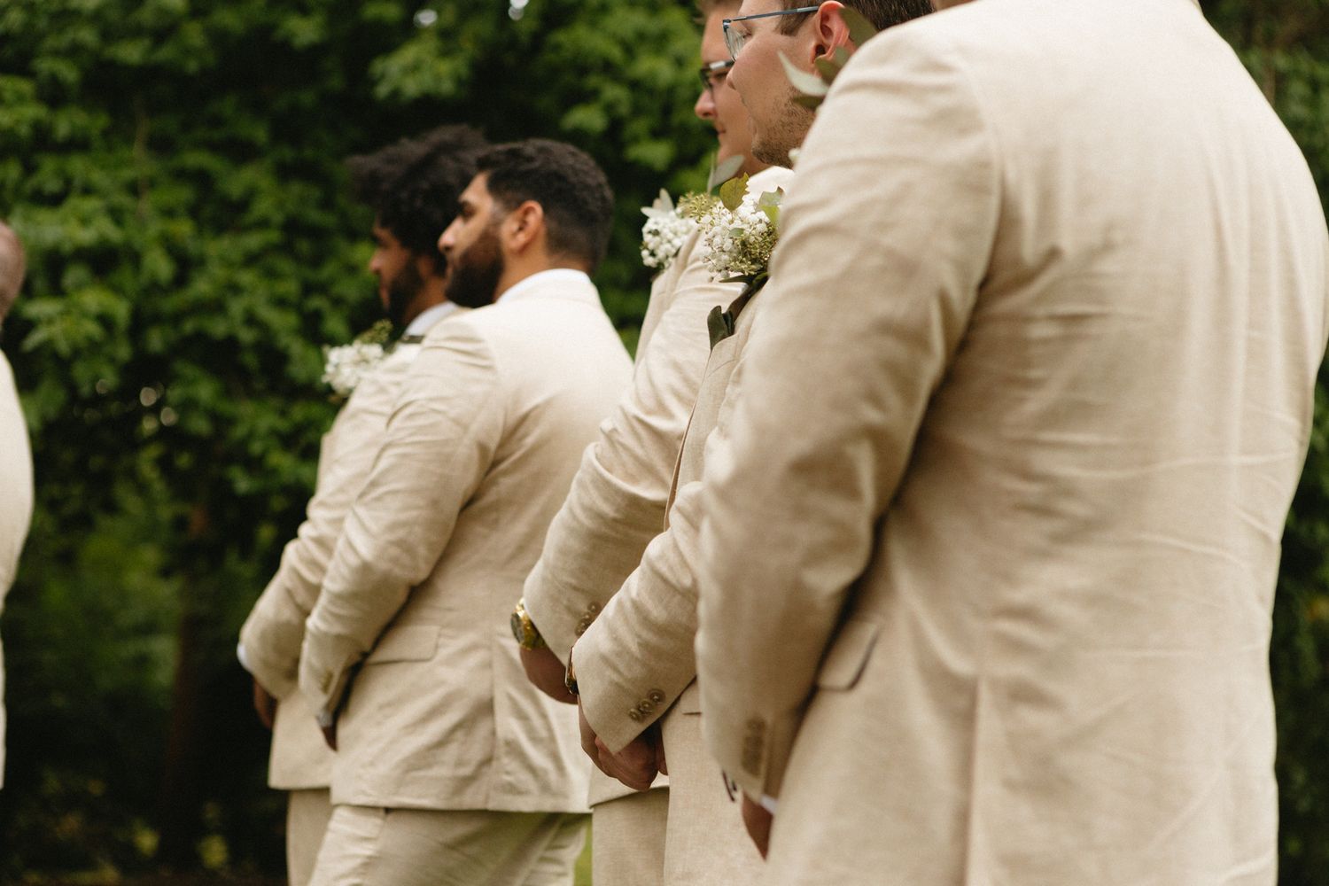 Groomsmen in cream colored suits stand in a line at an outdoor wedding ceremony with a boutonniere pinned to their lapels.