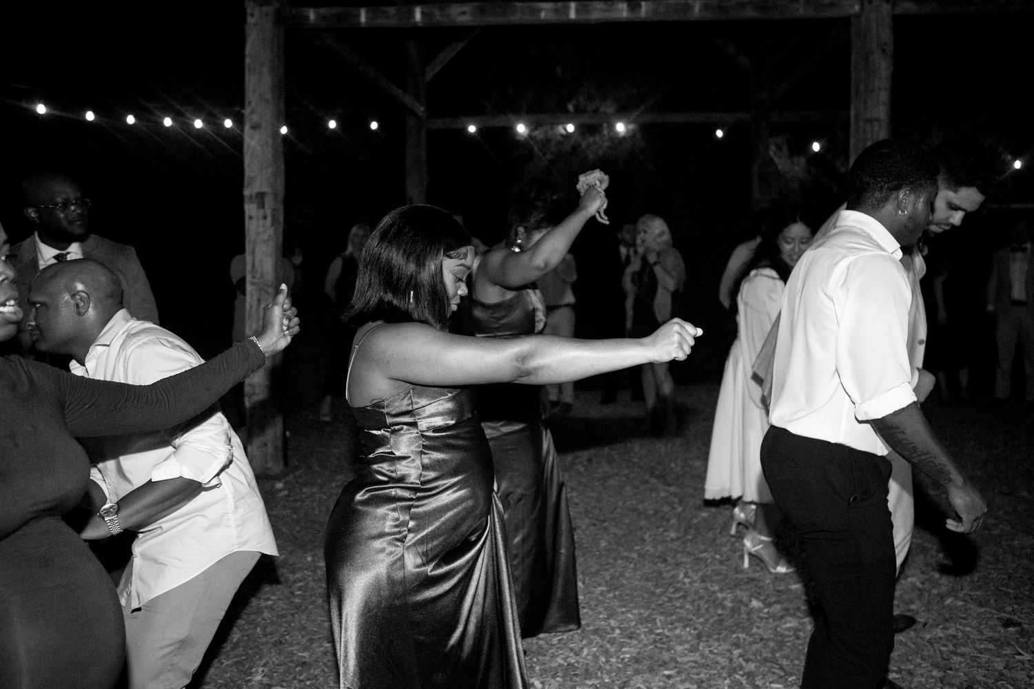 A black and white photo of couples dancing at a formal evening event with string lights overhead.