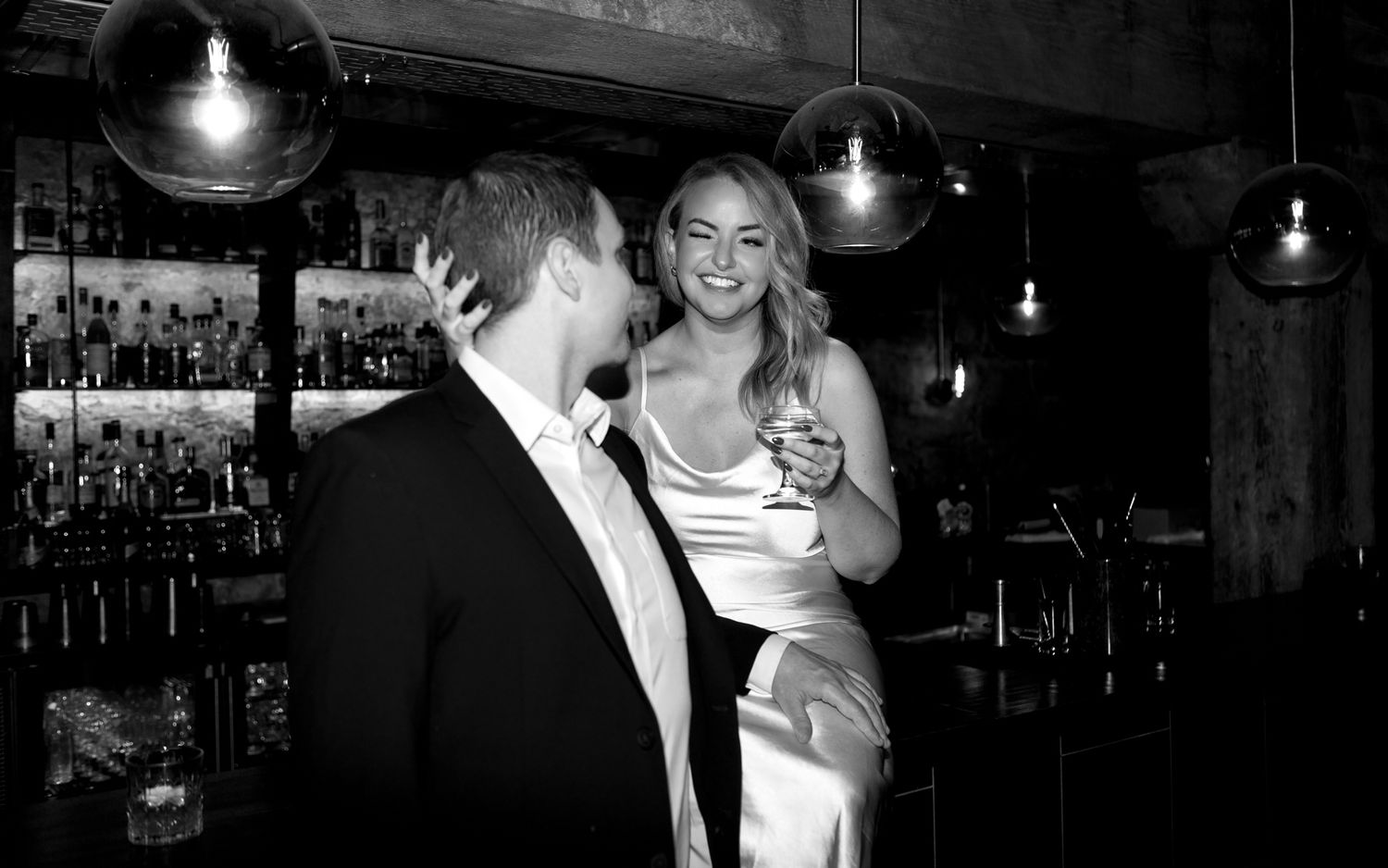 A couple shares a laugh together in elegant attire at a bar with pendant lighting and exposed brick walls.