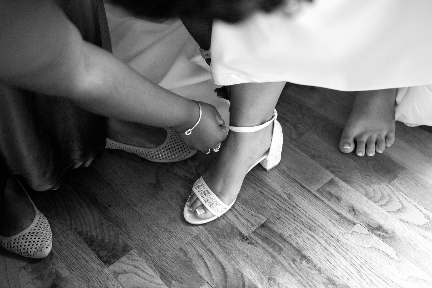 Black and white photo showing hands fastening a white ankle strap sandal on a hardwood floor.