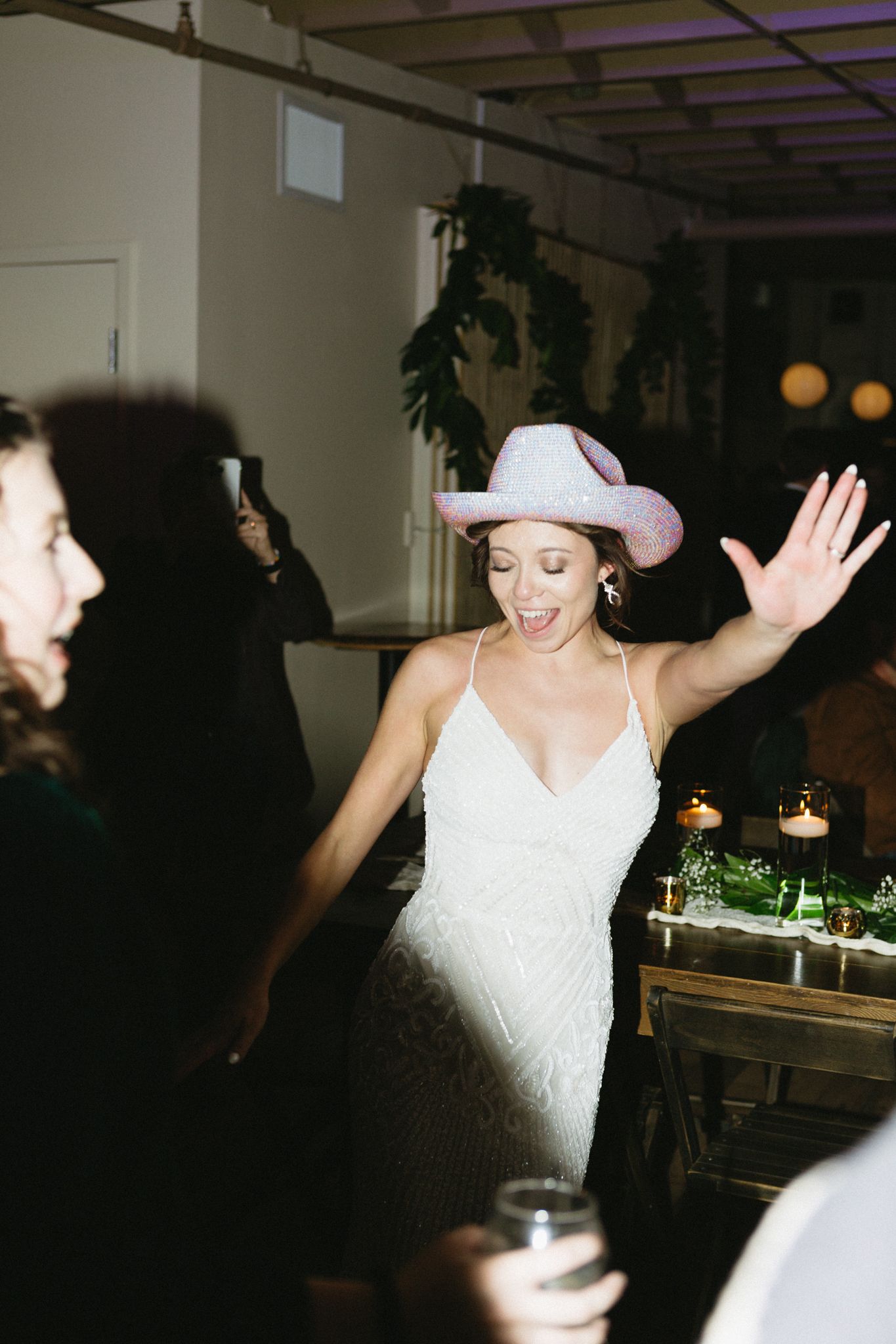 A bride in a white dress and pink cowboy hat dancing at a party with guests.