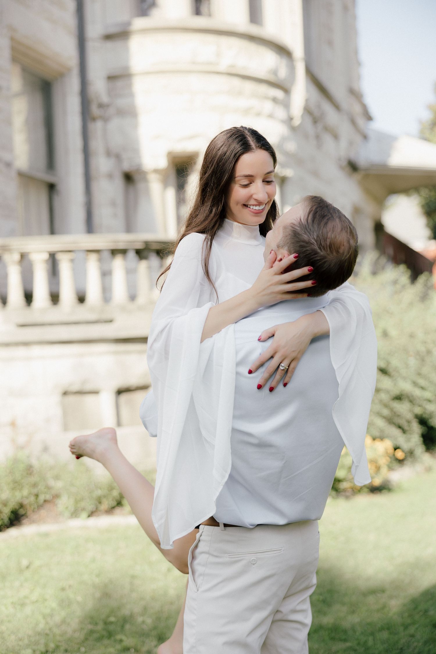 Couple in white embracing outdoors, man lifting woman near elegant stone building with balustrade.