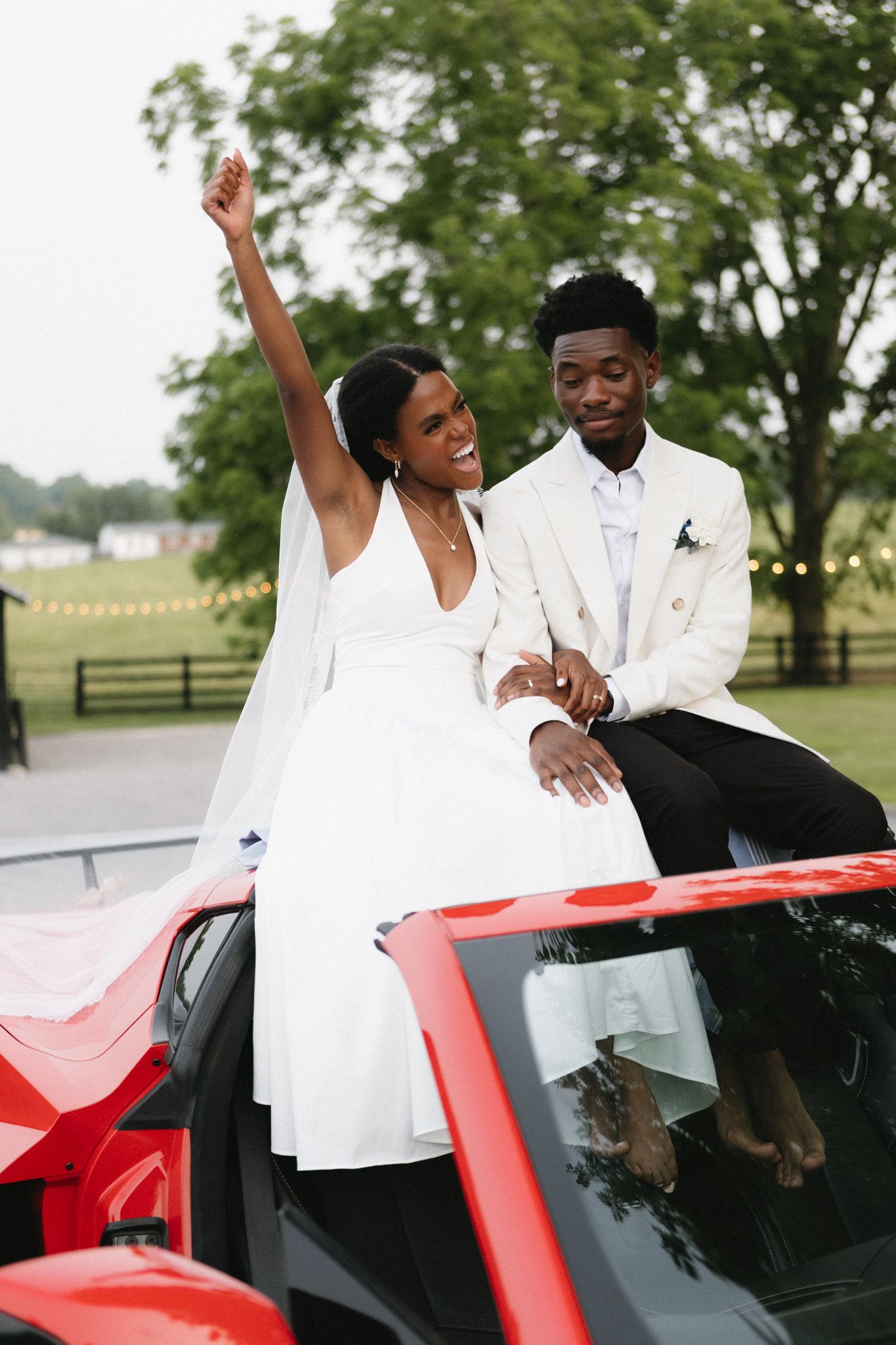 Happy newlyweds share playful moments while posing with a red sports car outdoors.