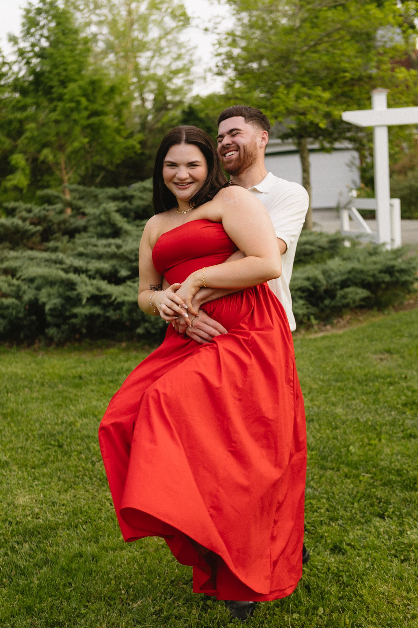 Romantic couple embracing in garden setting with red dress flowing in summer breeze.