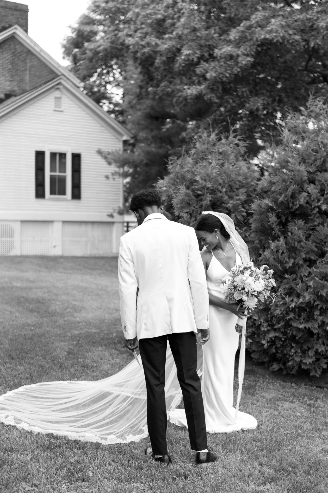 Black and white photo of couple near white house with long wedding veil trailing on grass.