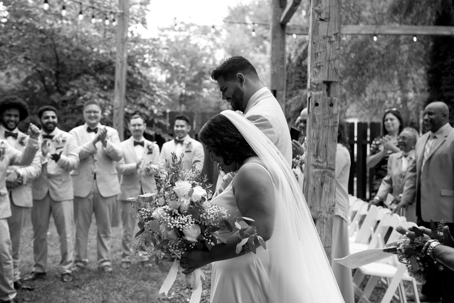 A black and white photograph captures a joyful wedding ceremony outdoors with groomsmen in light suits applauding.