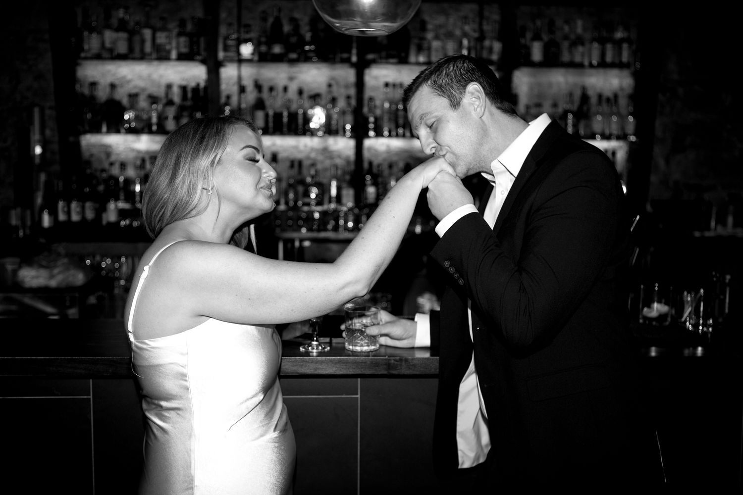 A romantic black and white moment at the bar as two people share an intimate gesture during a formal occasion.