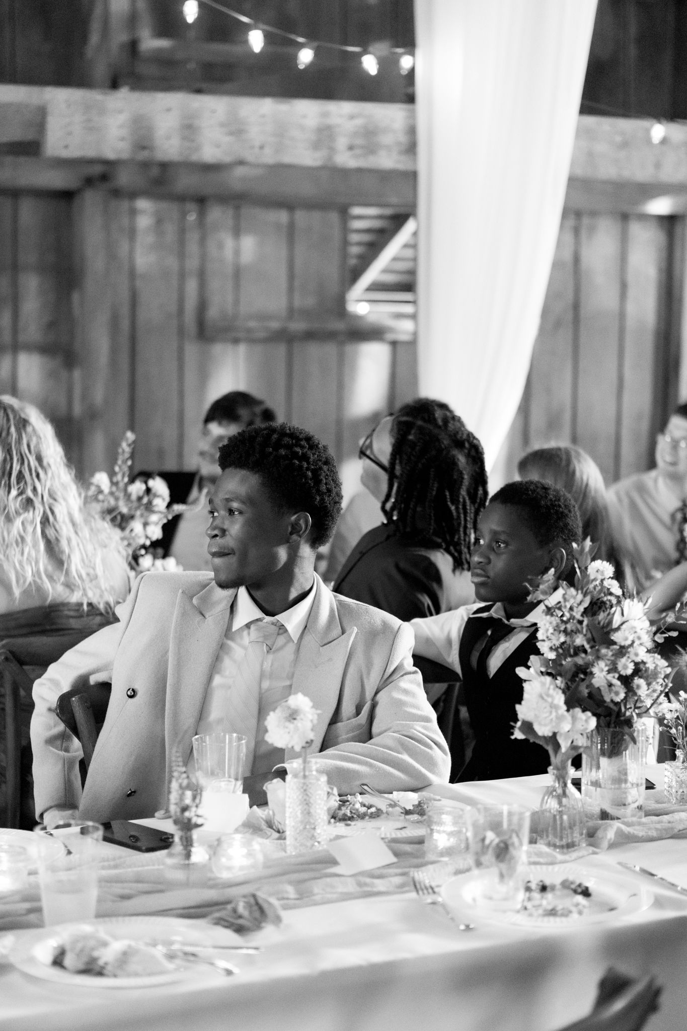 Black and white photo of wedding guests seated at a long reception table with floral centerpieces and candlelight.