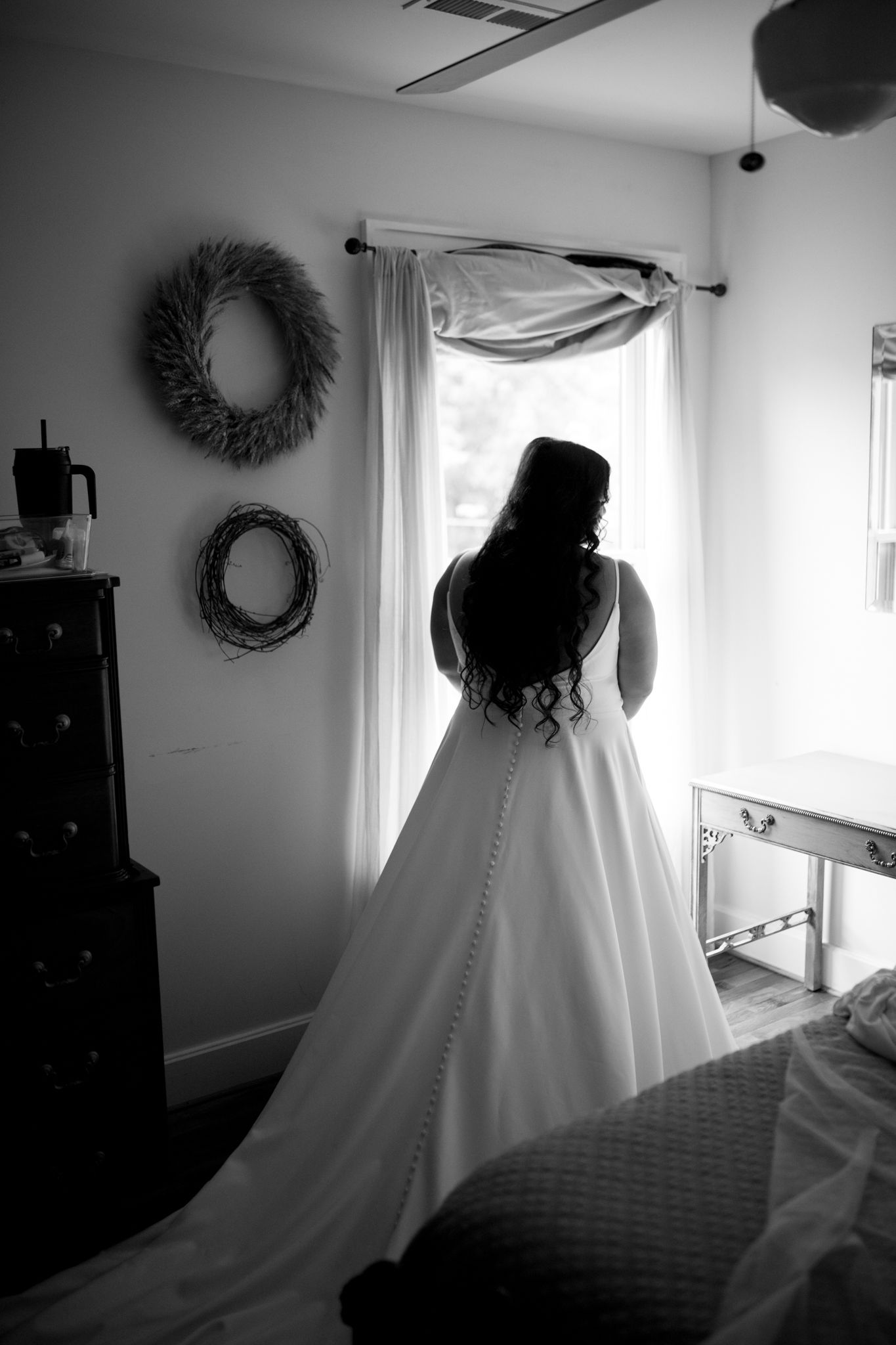A silhouette of a bride in a flowing white wedding gown stands by a window with decorative wreaths on the wall.