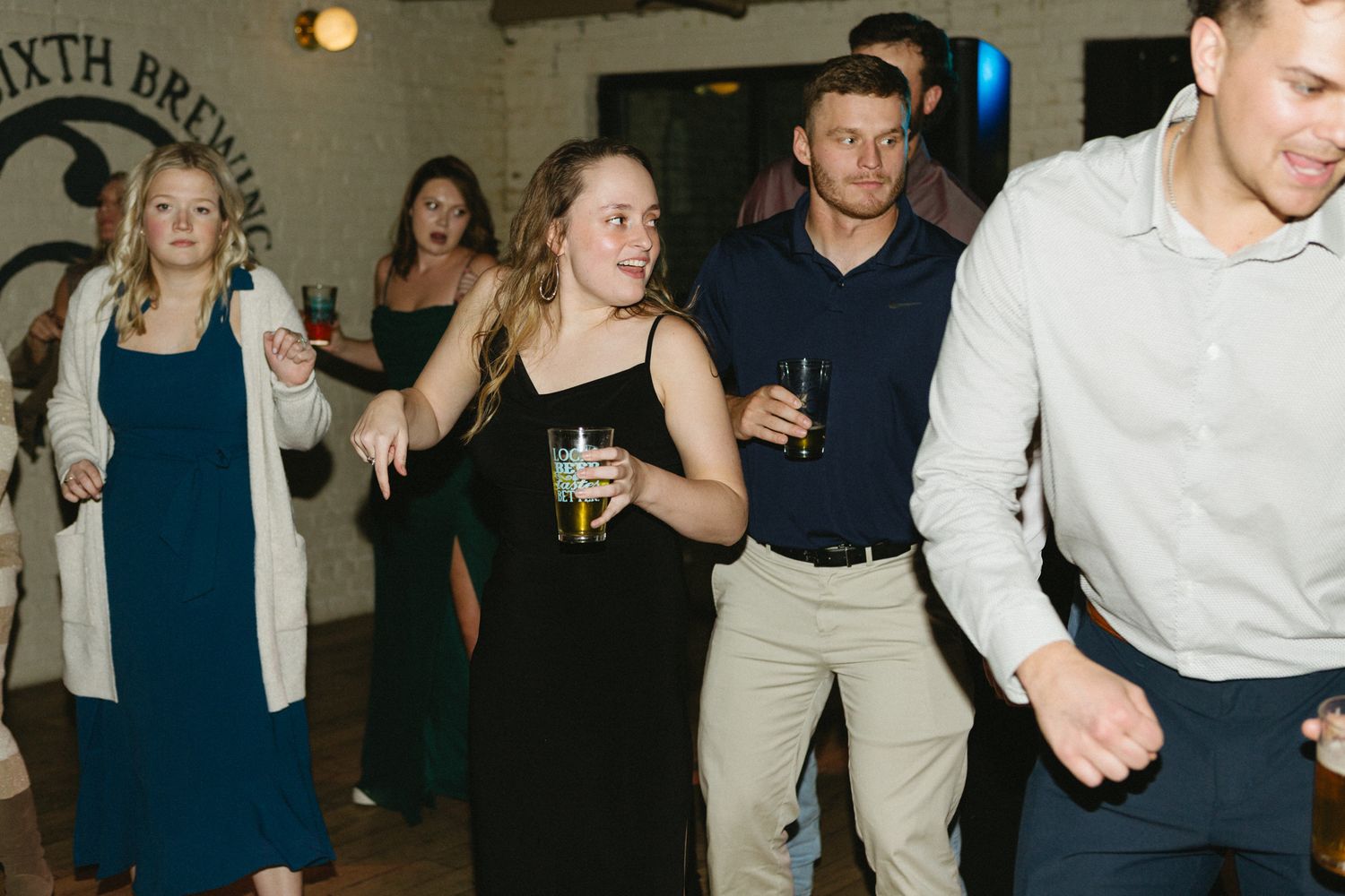 Party guests dance and celebrate with drinks at a lively indoor reception event.