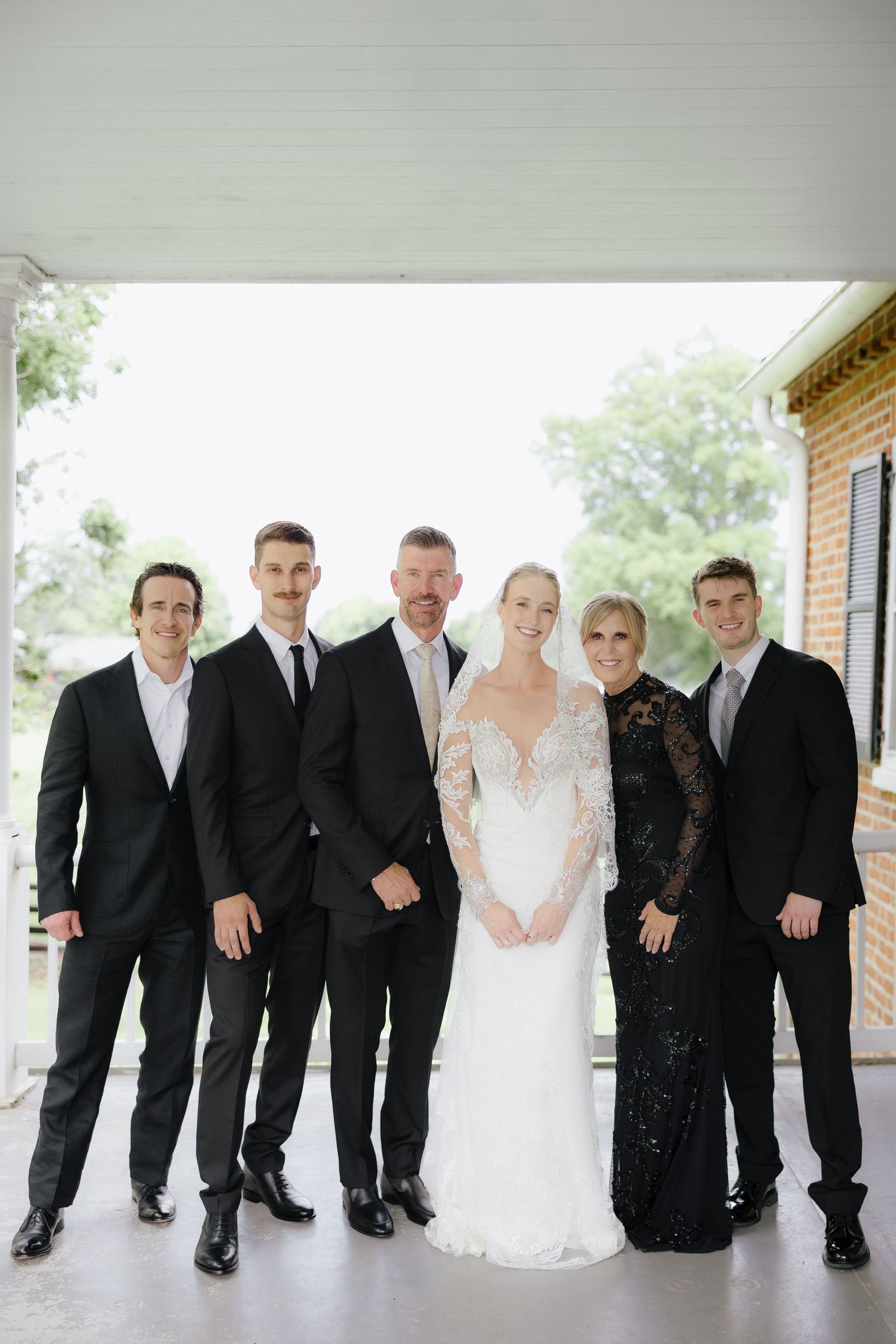 Bride in lace gown posing with family members dressed in black formal attire on porch.