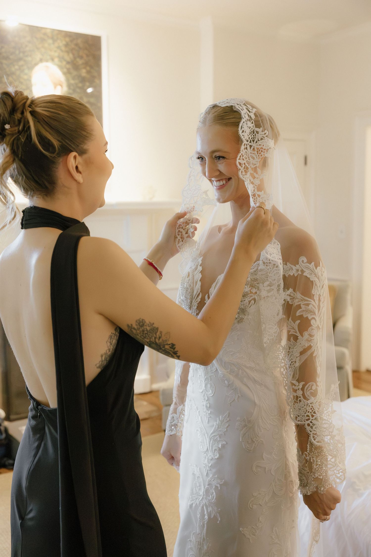 Bridesmaid in black dress adjusts lace veil on smiling bride in elegant lace wedding gown indoors.