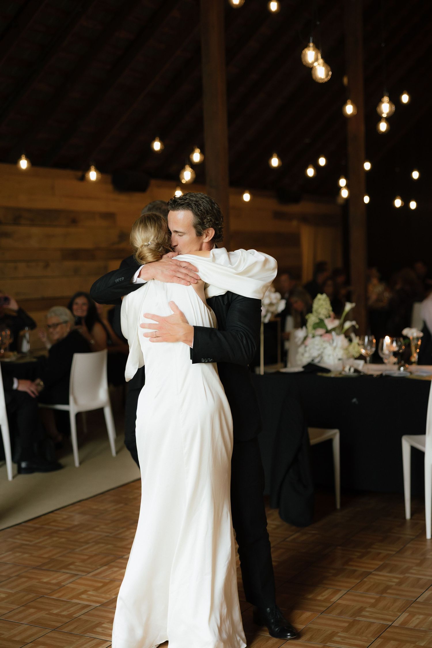 Bride and groom share romantic first dance embrace in rustic barn venue with warm Edison bulb string lights.