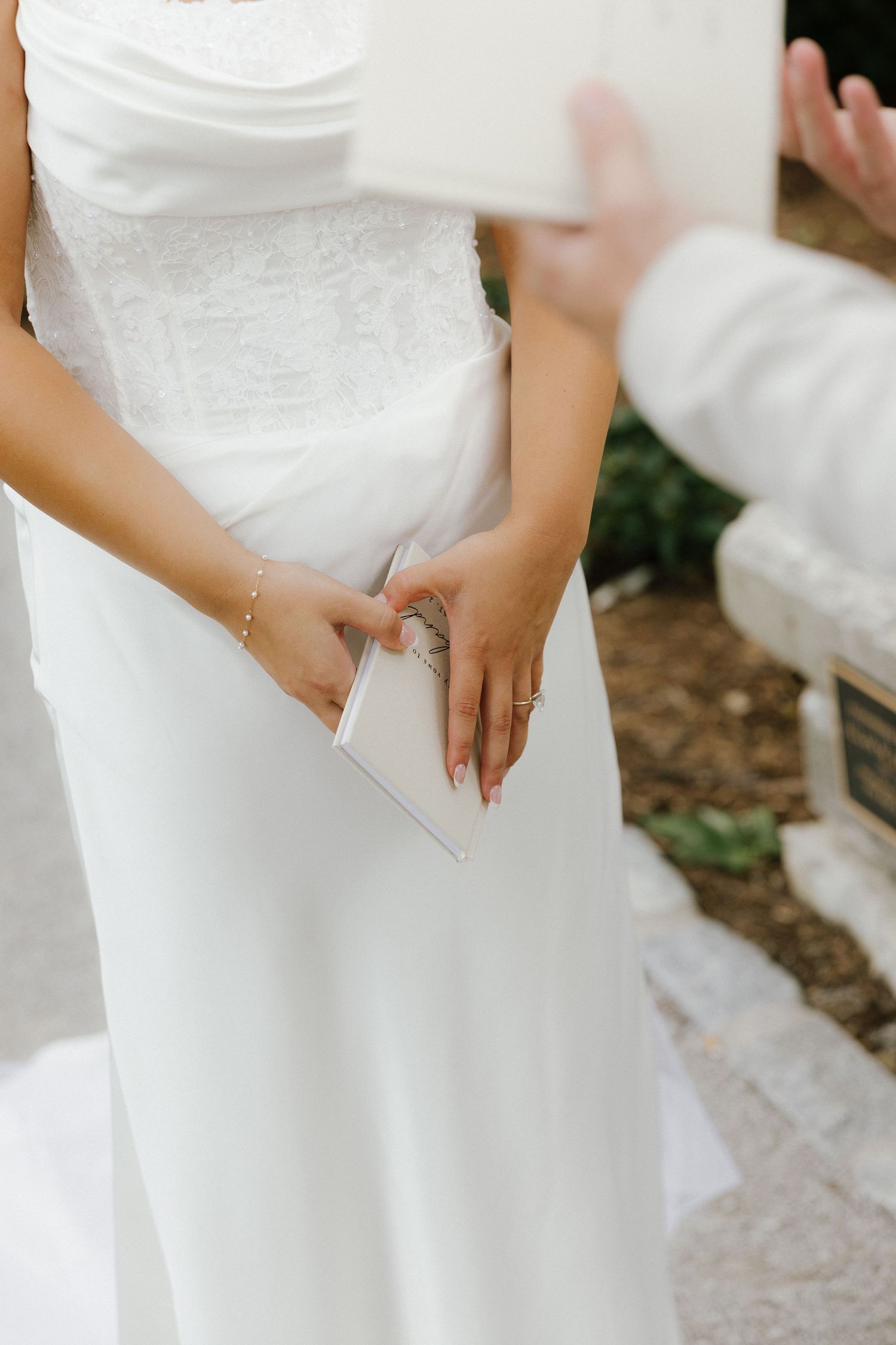 Bride in white lace gown holding vow booklet during outdoor wedding ceremony.