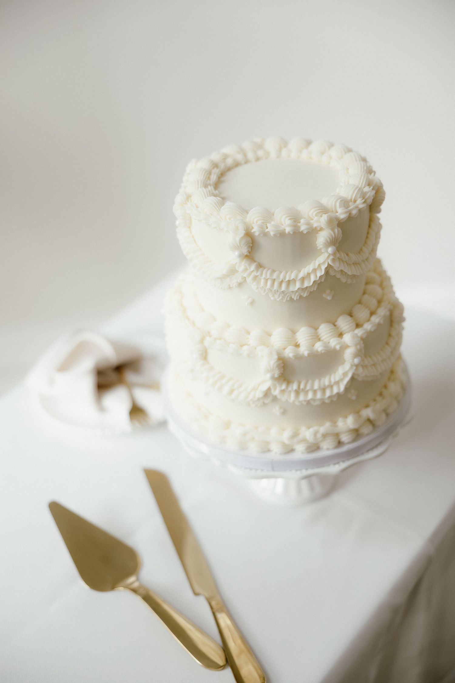 Elegant three-tier white wedding cake with pearl details on cake stand with gold serving utensils.