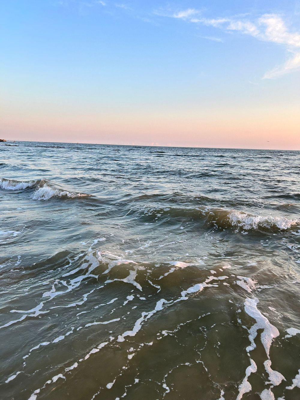 Gentle ocean waves washing ashore at sunset with soft pink sky and blue horizon over calm sea.