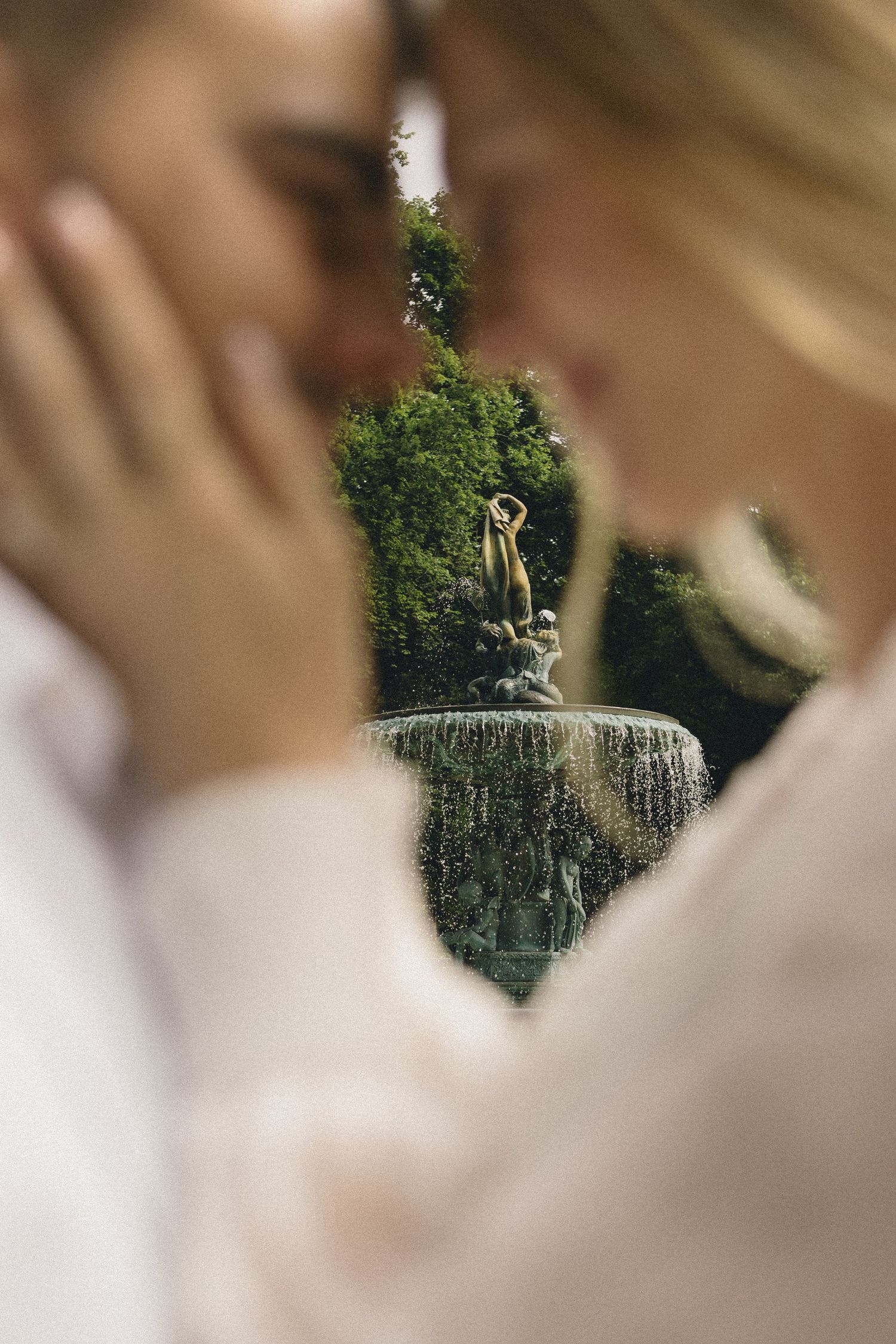 Romantic couple almost kissing with a lush green fountain statue beautifully blurred in the background.