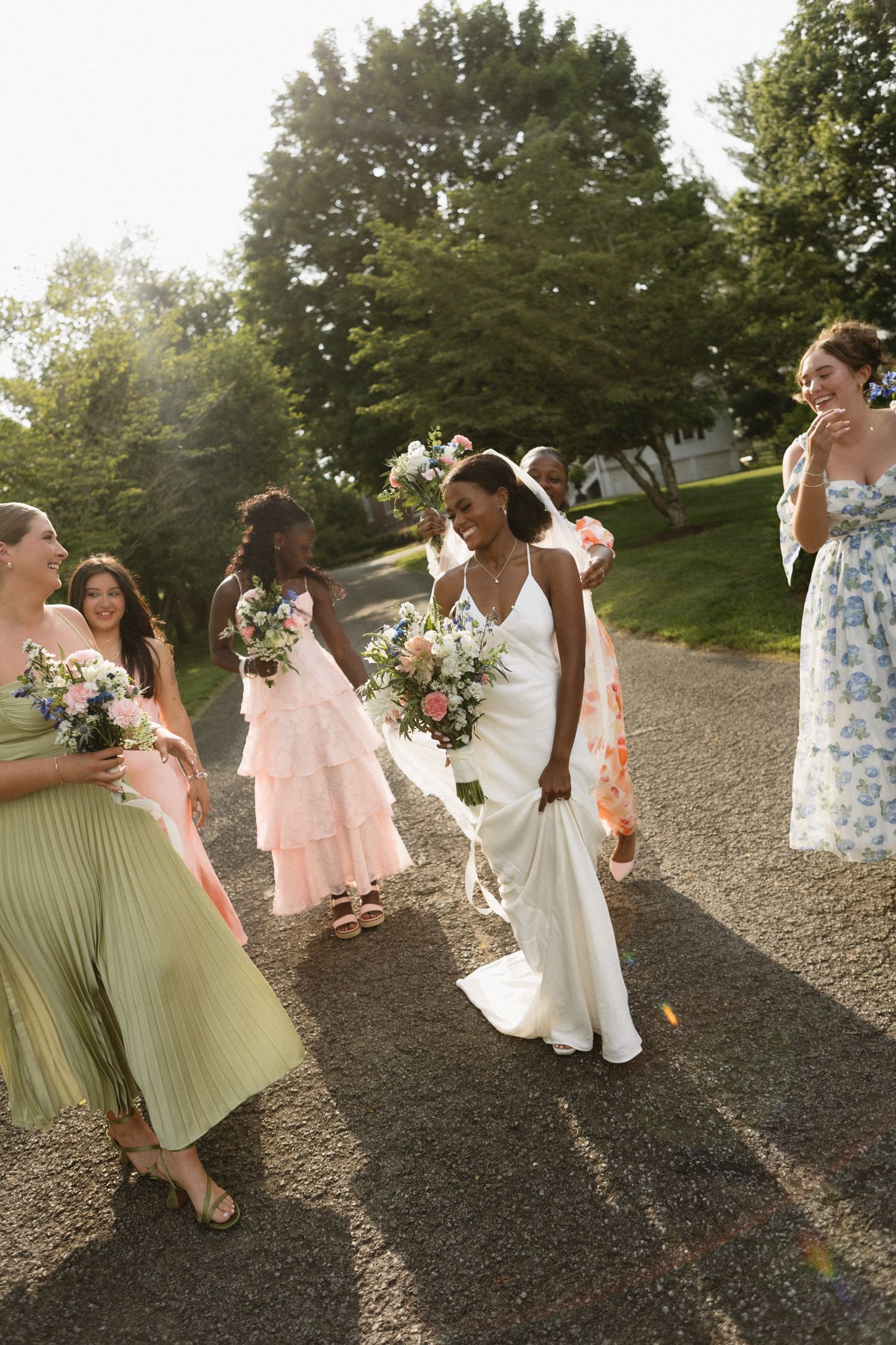 Bride in white gown walks with bridesmaids in colorful dresses, holding bouquets outdoors on a sunny day.