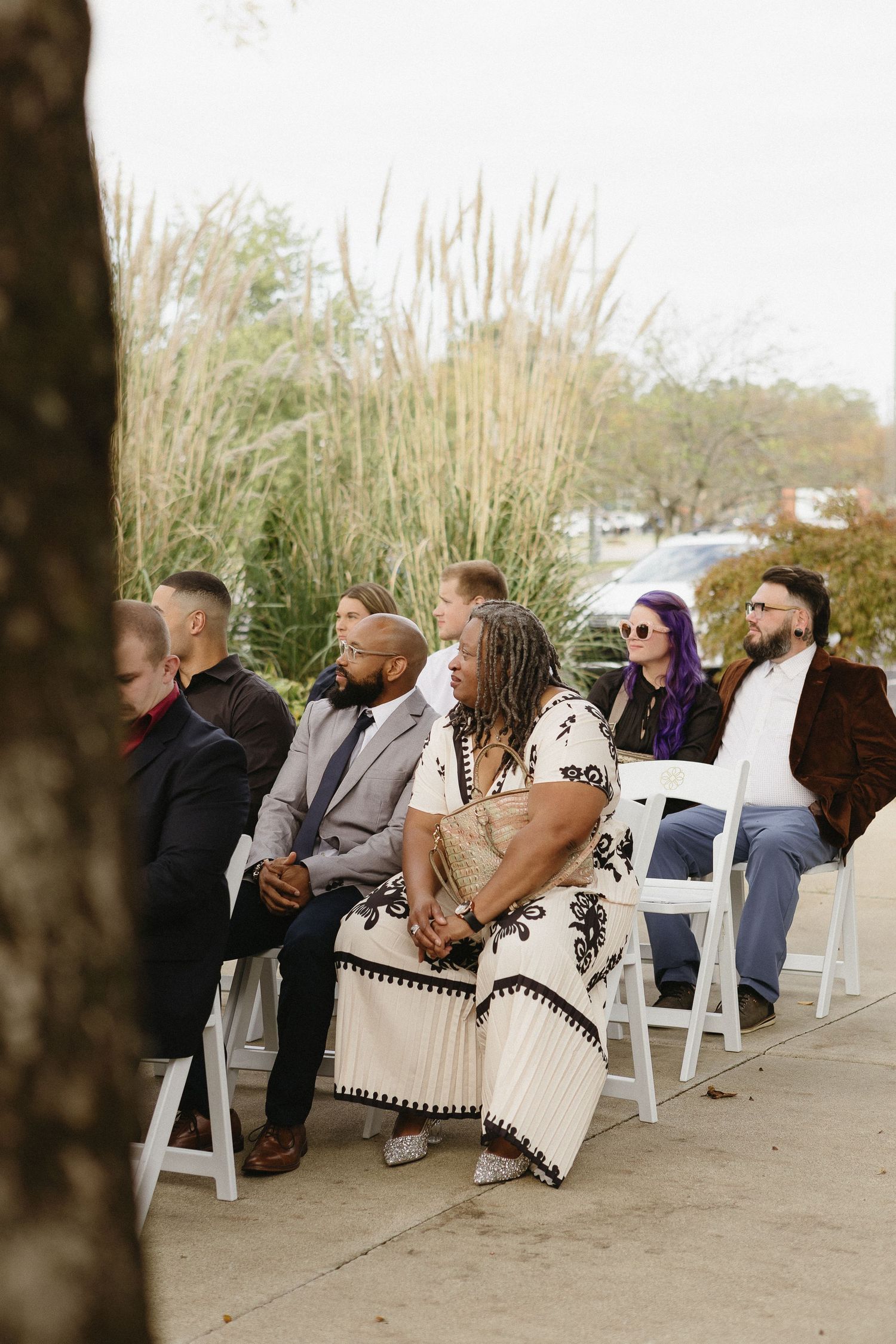 Wedding guests seated on white chairs outdoors with tall grass in the background.
