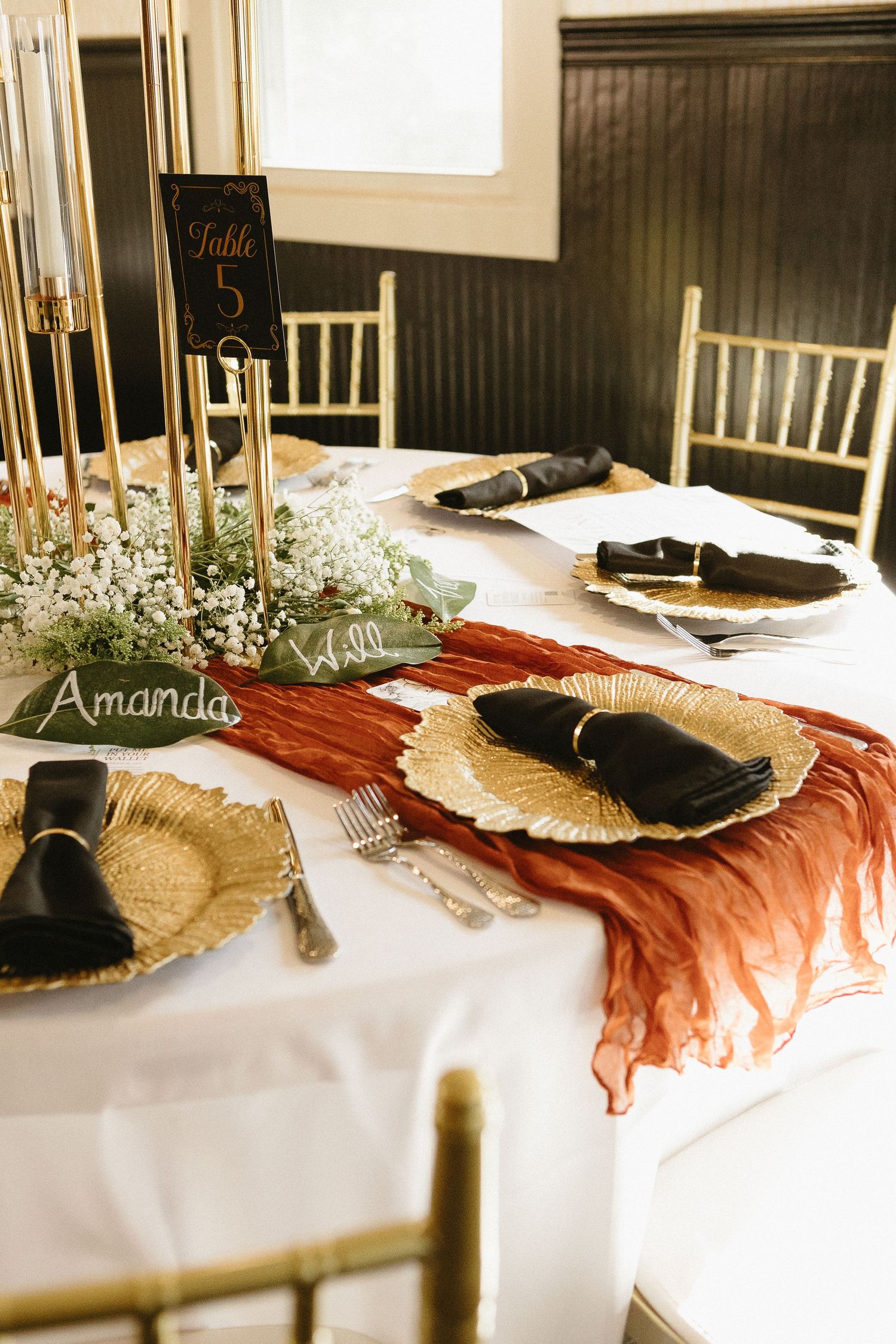 Wedding reception table settings with gold chargers, burgundy accents, and name cards.