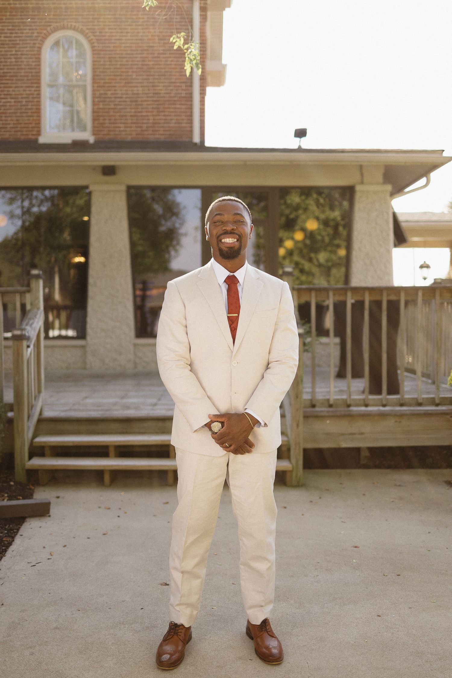 Groom in cream suit and red tie poses outside brick building entrance.
