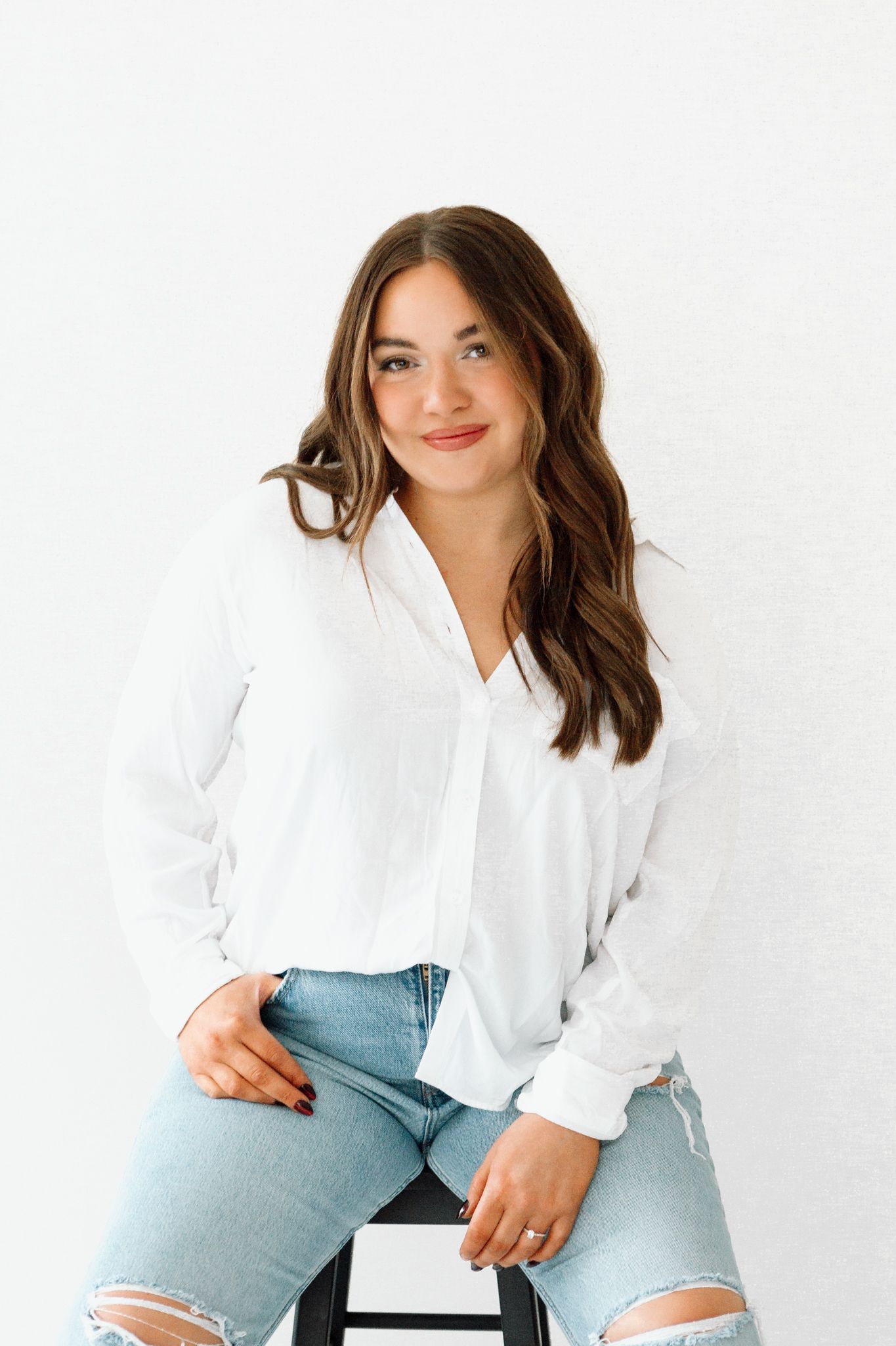 Young woman in white button-up shirt and ripped jeans sitting on stool against white background.
