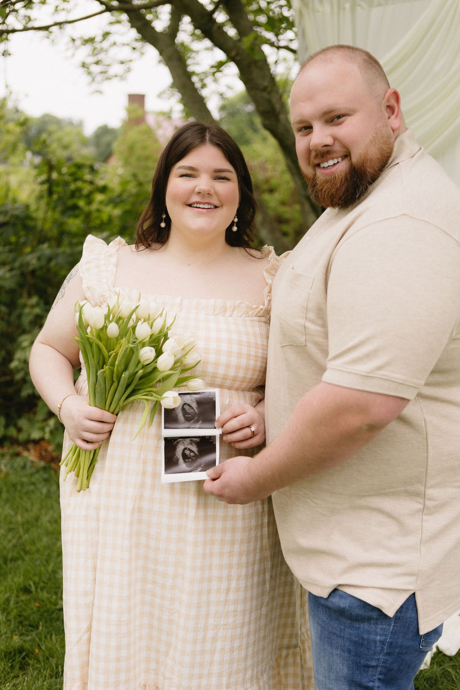 Happy couple outdoors holding ultrasound photos and white tulips, announcing pregnancy in a garden setting.