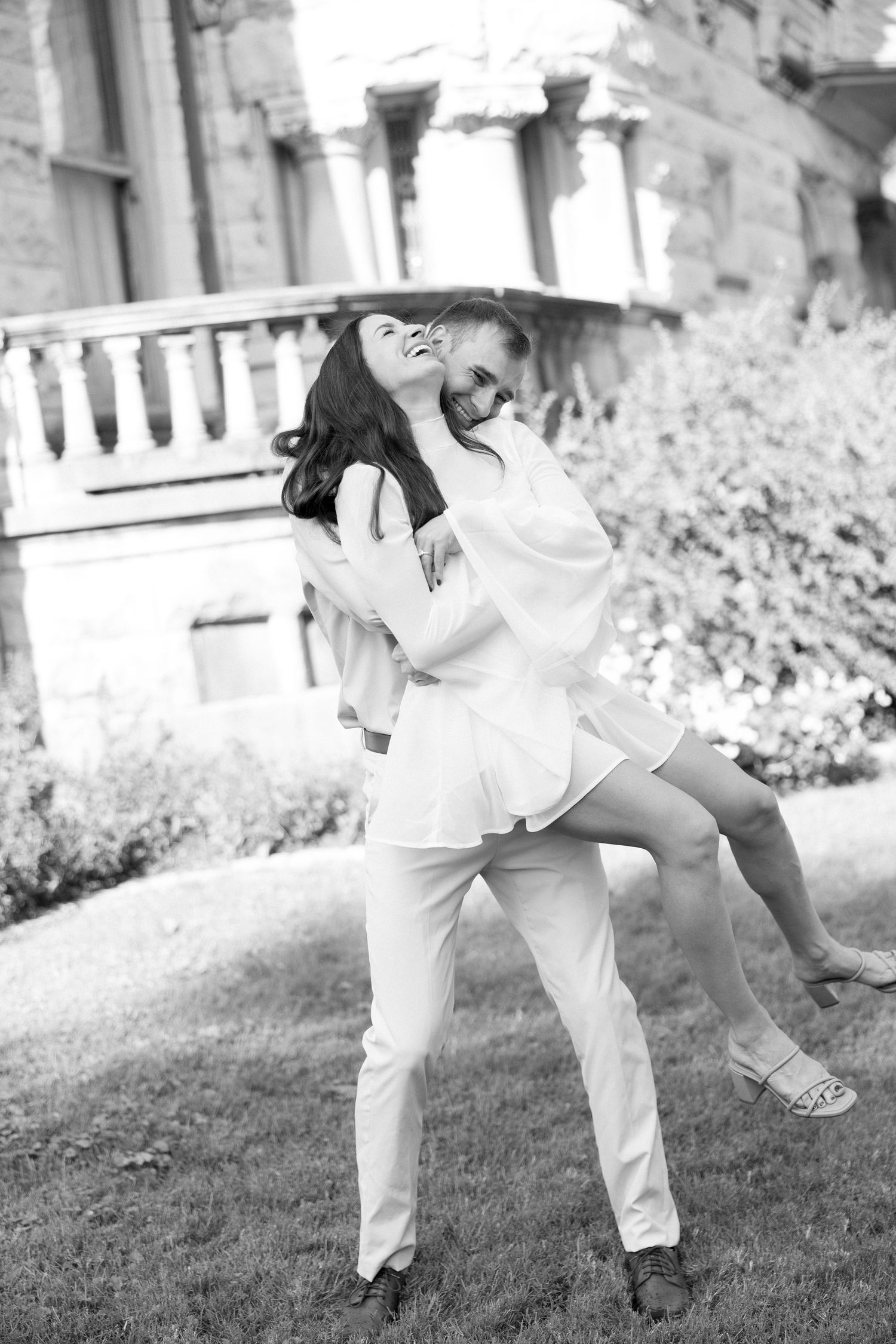 Black and white photo of a joyful couple dancing outdoors, man lifting woman in white dress.