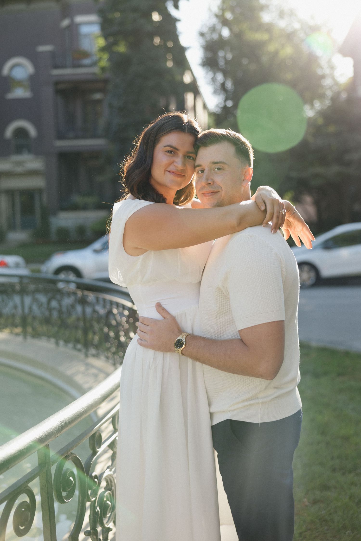 A couple embraces near a decorative railing outdoors, woman in white dress hugging man in cream shirt, golden sunlight behind them.