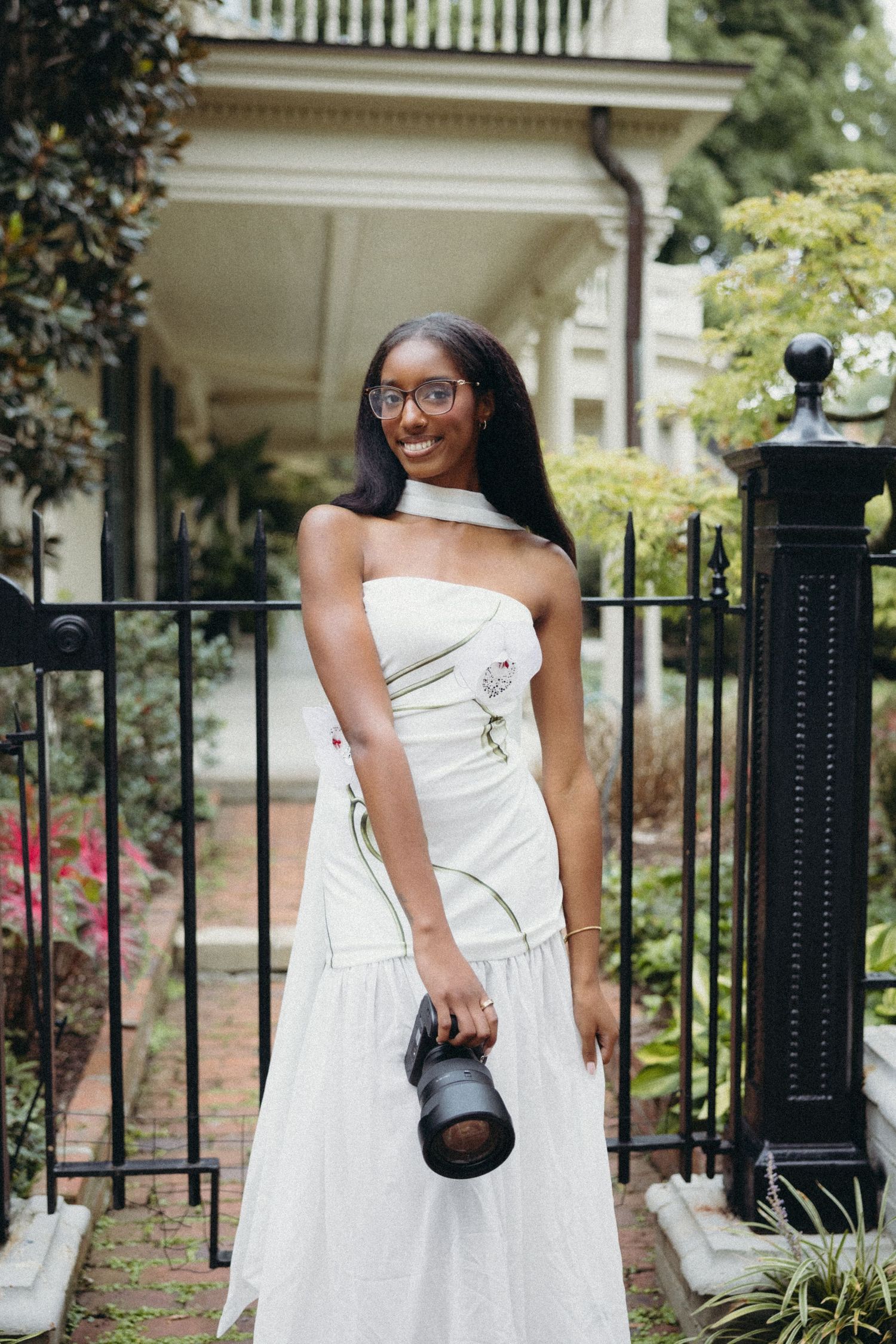 Young Black woman in white strapless gown holding camera, smiling outdoors near iron gate and historic home.