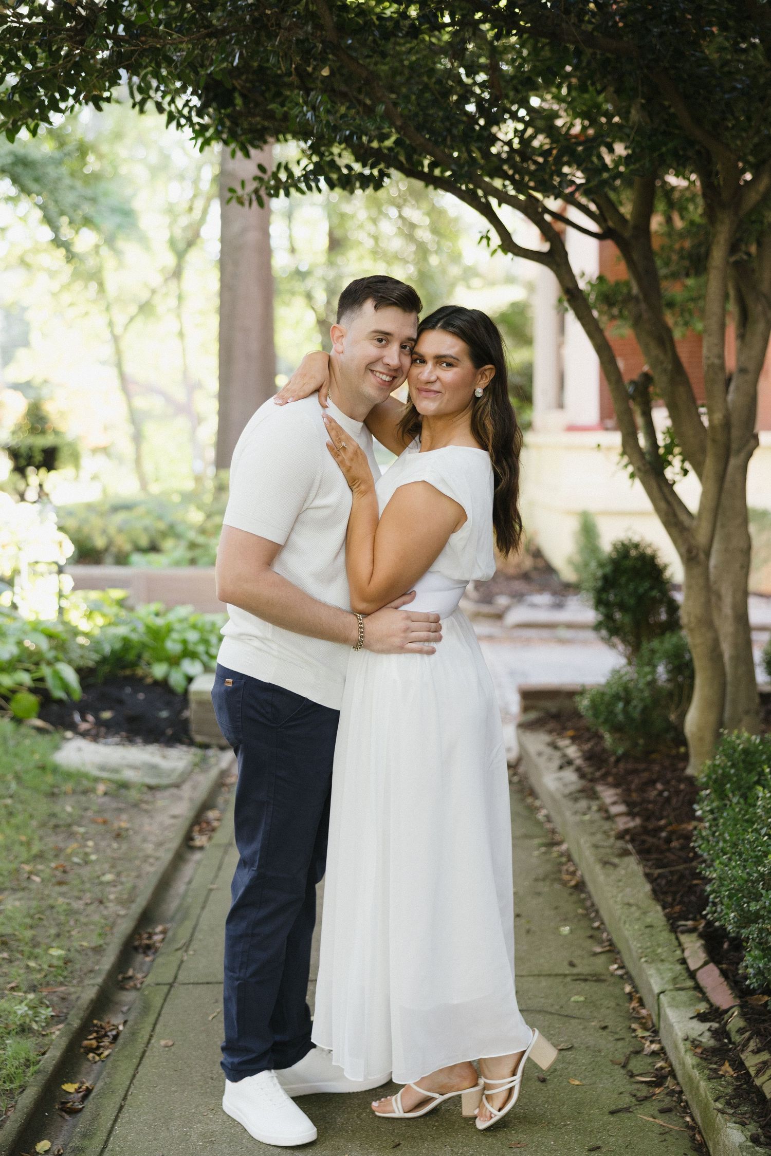 Happy couple in white outfits share a romantic embrace on a shaded garden walkway.