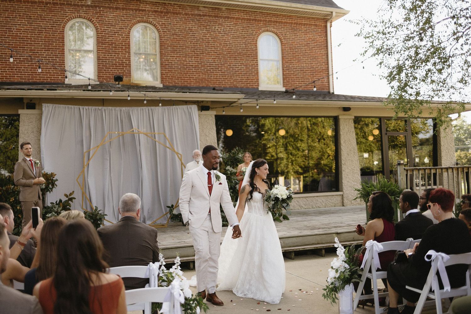 Wedding ceremony setup outside a brick building with white chairs and floral arrangements.