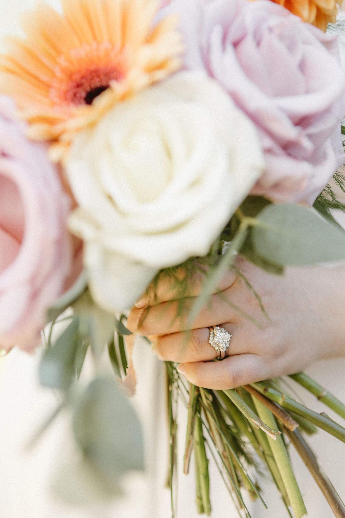 A close-up view of a delicate wedding bouquet featuring peach gerbera daisies and white roses with a sparkling engagement ring.