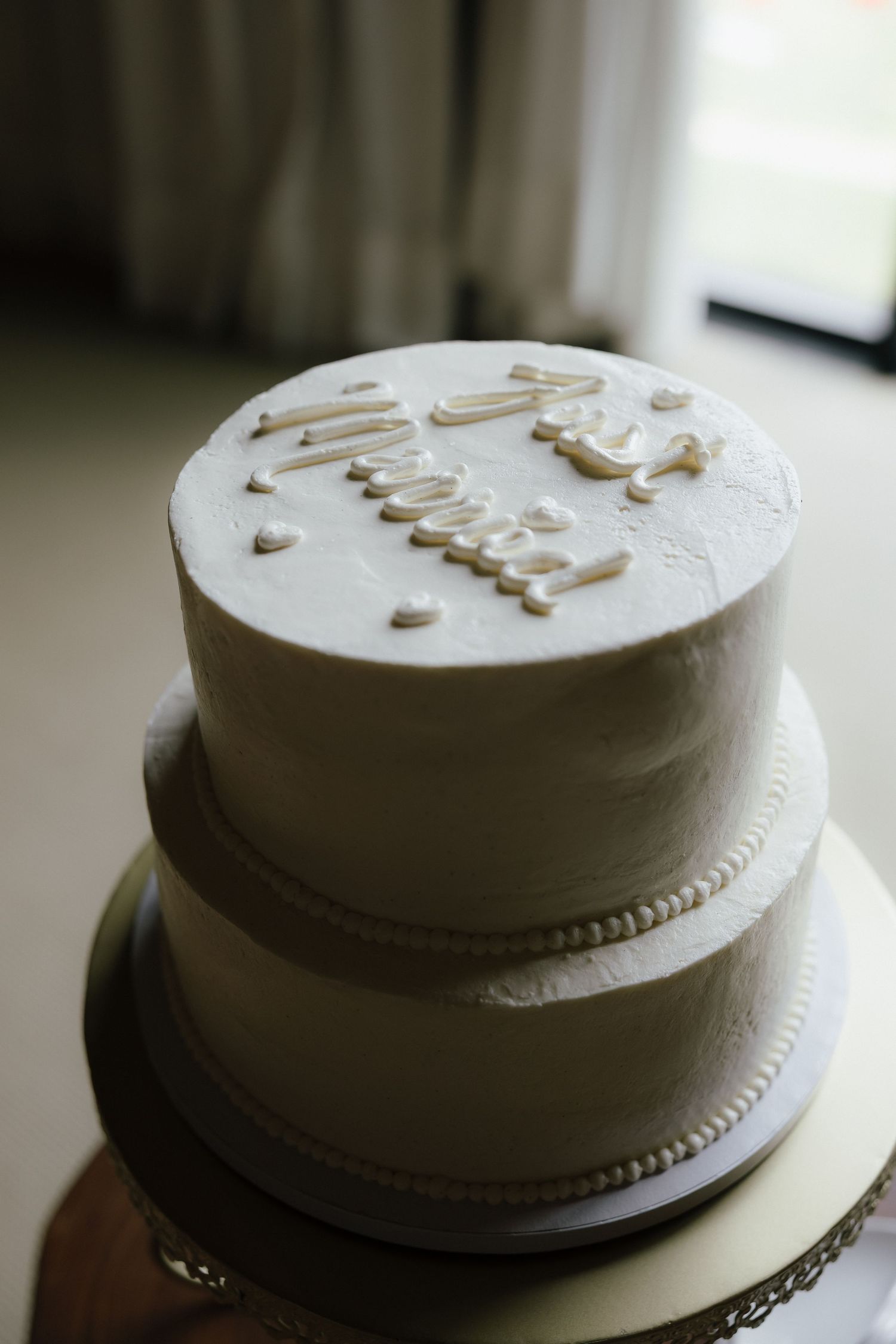 Two-tier white wedding cake with elegant script lettering on gold cake stand.