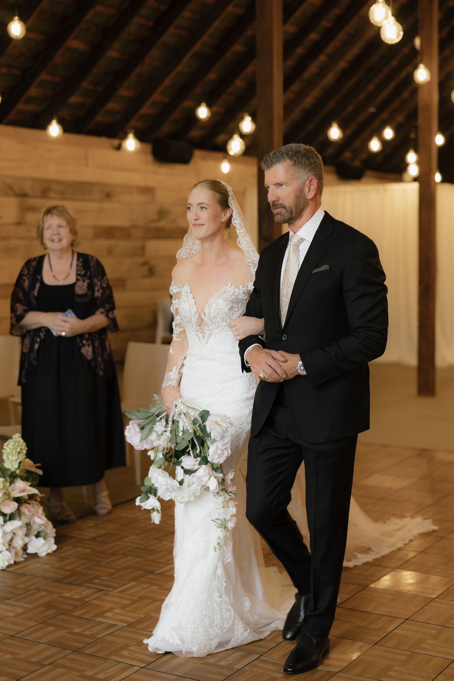 Bride in lace gown walks down aisle with father in black suit at rustic barn wedding ceremony.