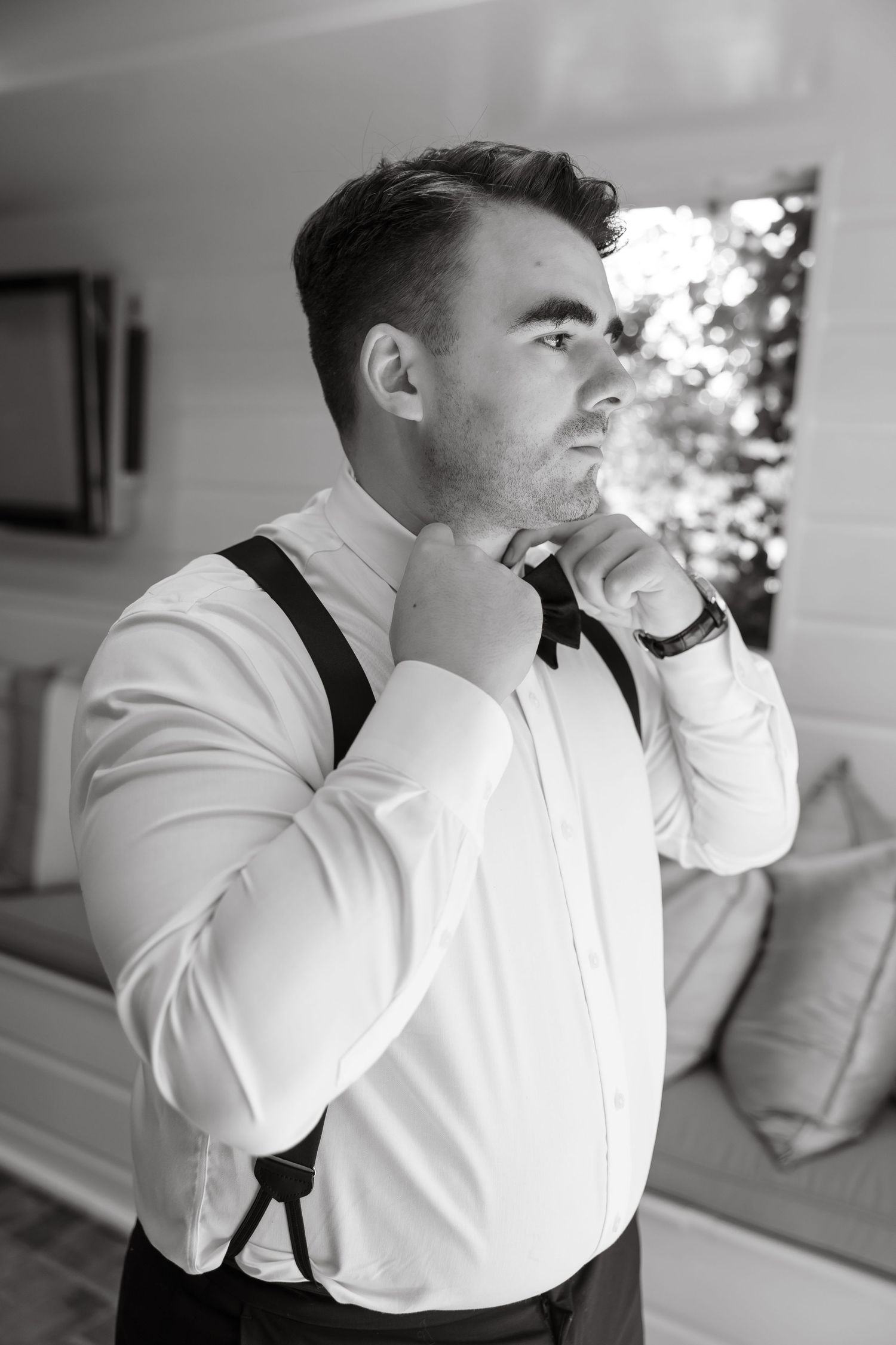 Groom adjusting bow tie wearing white shirt and suspenders in black and white wedding preparation photo.