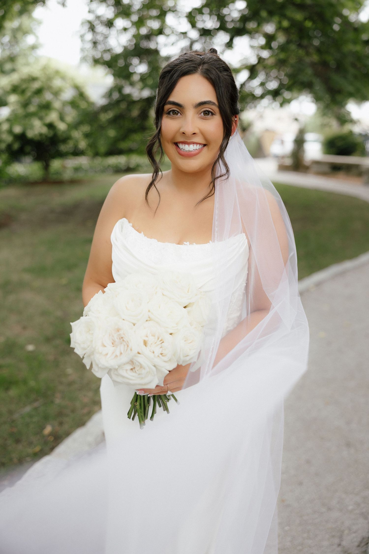 Smiling bride in white strapless gown holding white peony bouquet with veil outdoors.