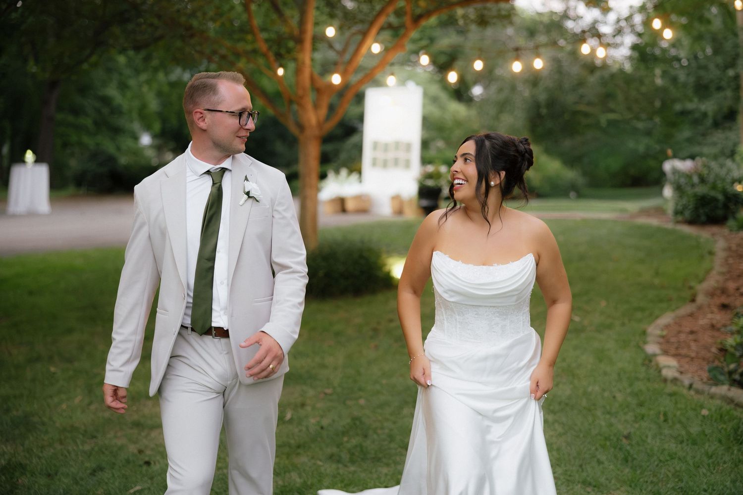 Bride and groom walk together laughing in a lush garden with string lights.