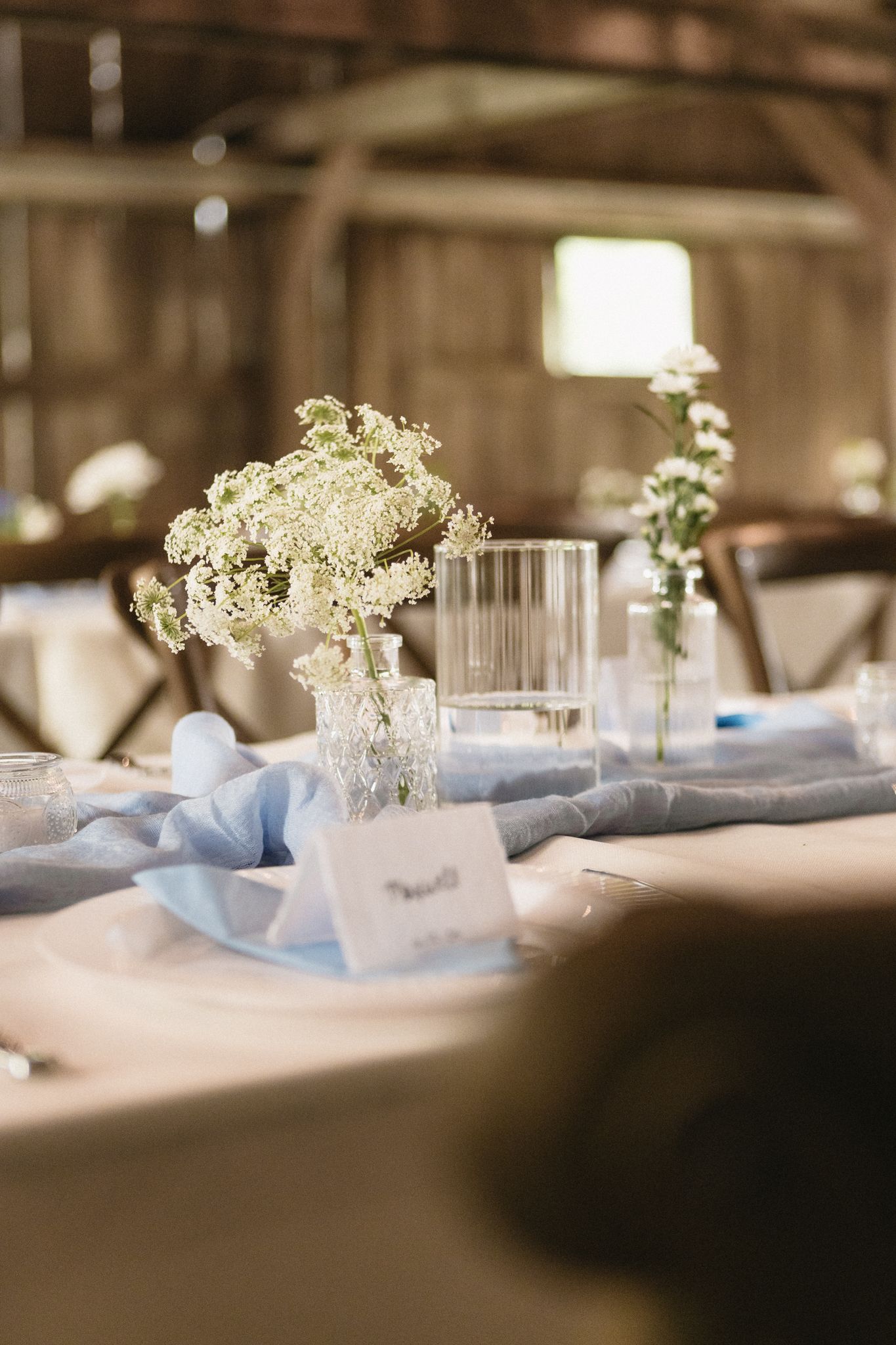 White floral centerpieces in glass vases decorate elegant wedding reception tables with soft blue accents.