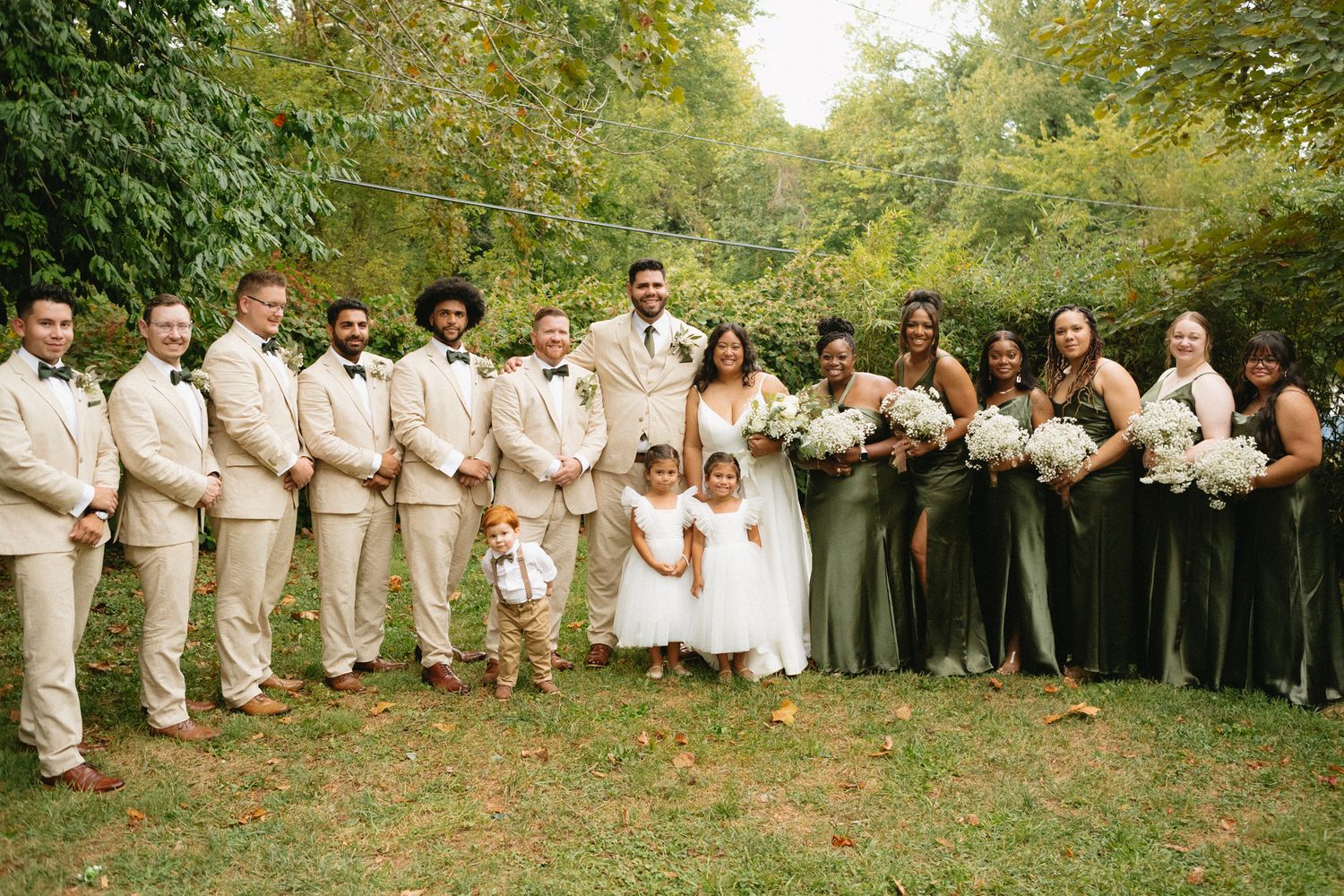 Wedding party in beige suits and sage green dresses poses together on autumn grounds during outdoor celebration.